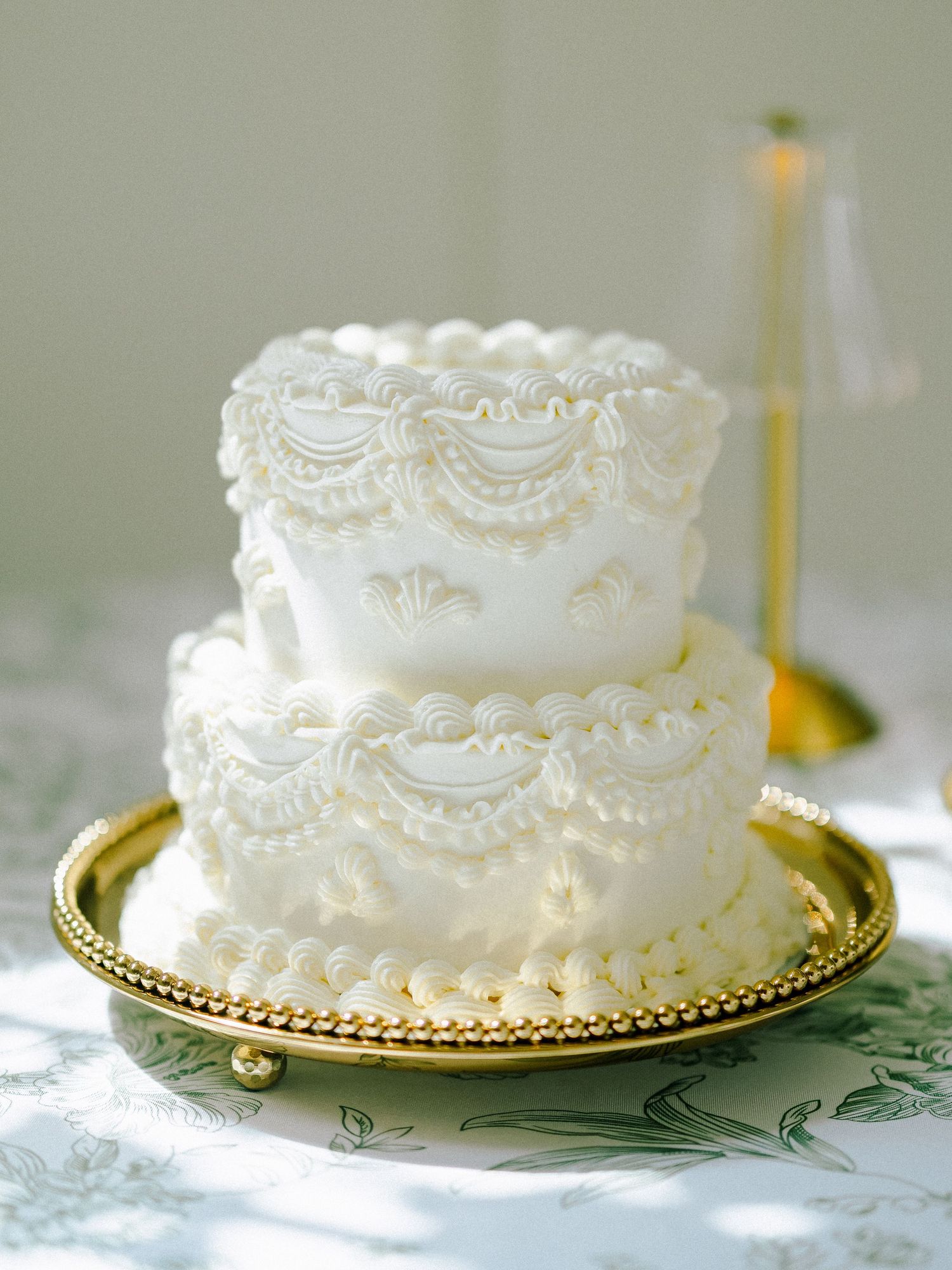 Elegant two-tier white wedding cake with intricate lace piping on a gold beaded cake stand.