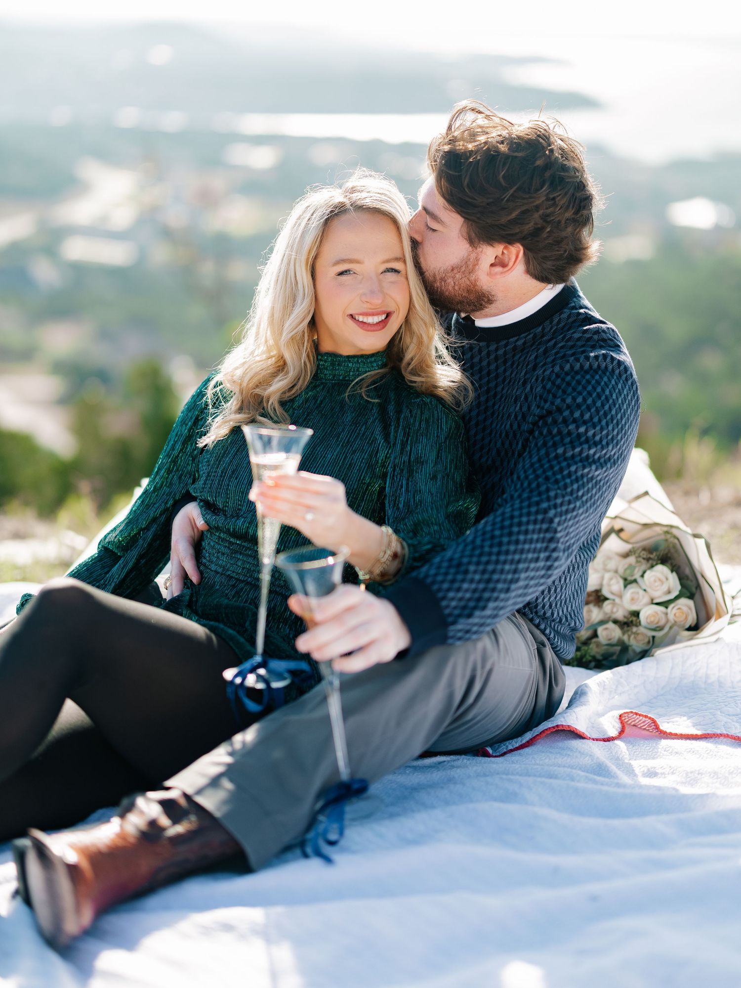 Happy couple sitting on a blanket outdoors, sharing champagne and a romantic kiss.