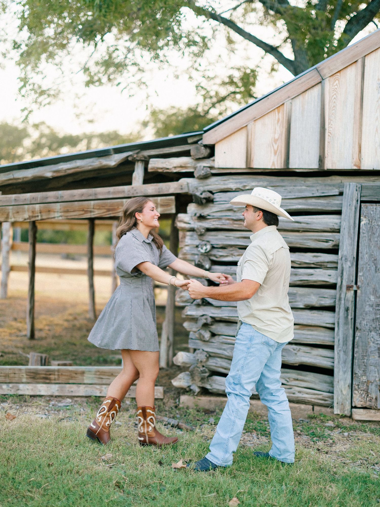Couple dancing and holding hands in front of a rustic log barn wearing cowboy boots and casual country attire.