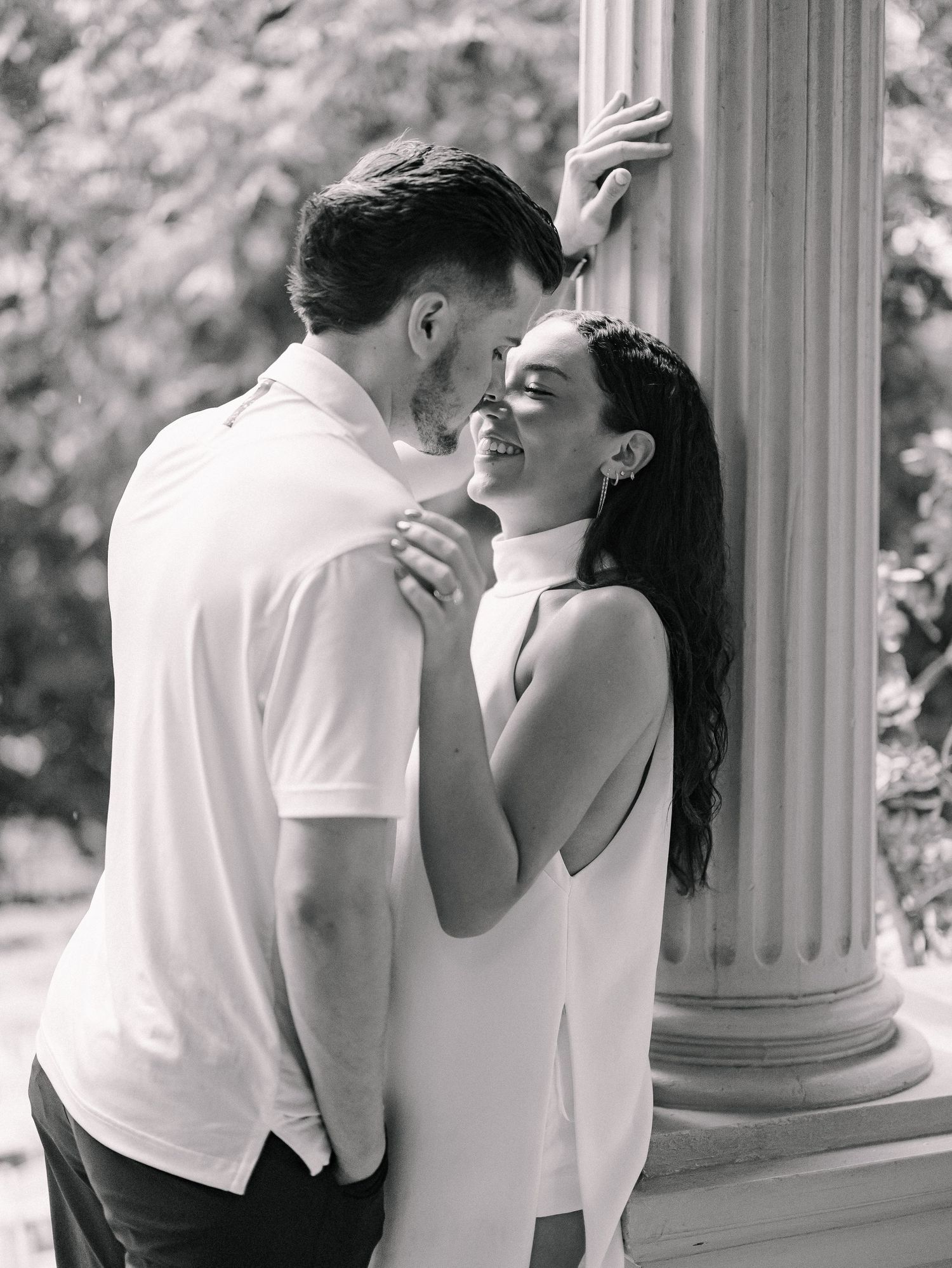 Black and white photo of a smiling couple sharing a romantic moment near a classical column outdoors.