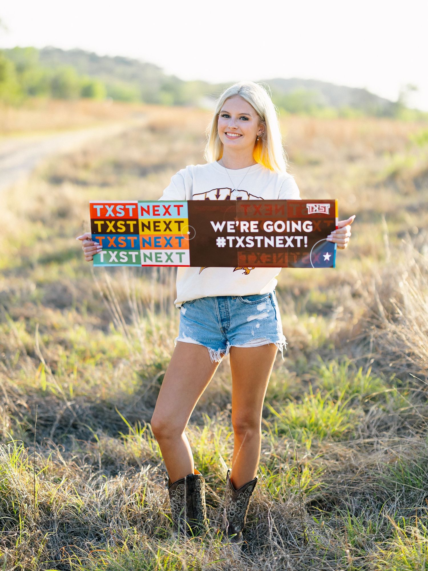 Smiling blonde woman holding TXST Next signs in a field, wearing denim shorts, cowboy boots, and a white t-shirt.
