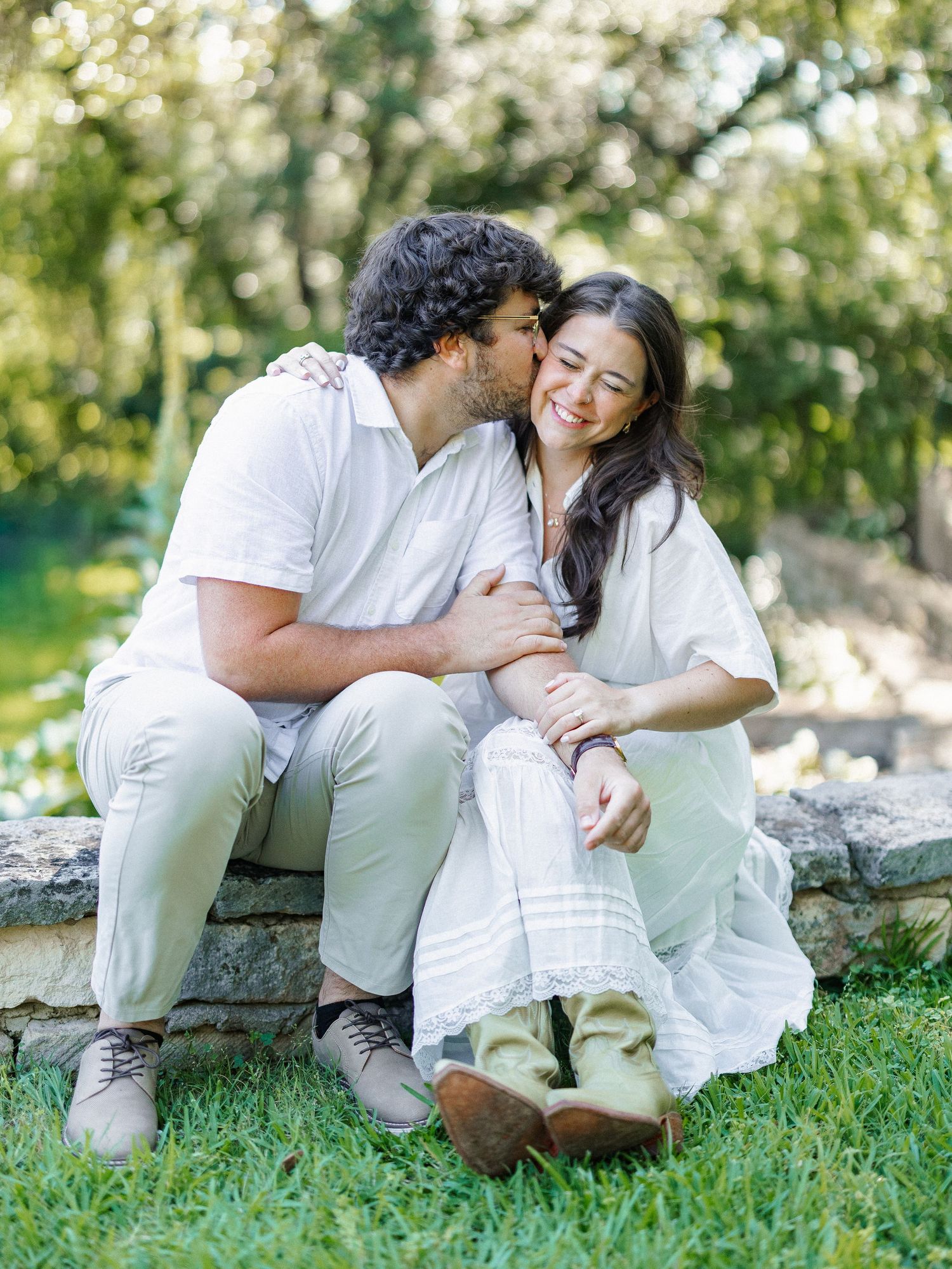 A couple sits on a stone ledge outdoors, man kissing woman on cheek, both wearing white in a lush green garden.