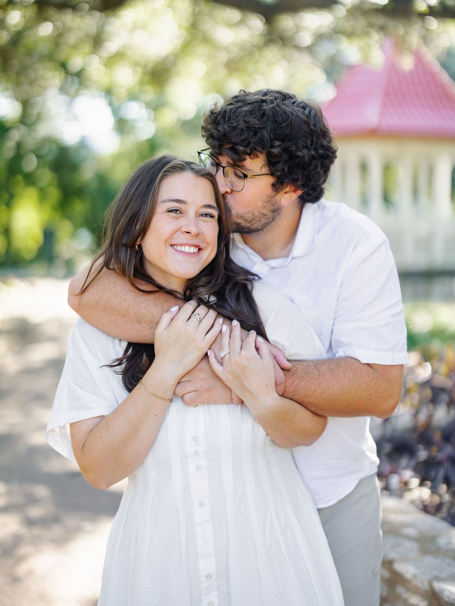 A couple embraces outdoors, woman in white dress smiling as man kisses her cheek near a pink gazebo.
