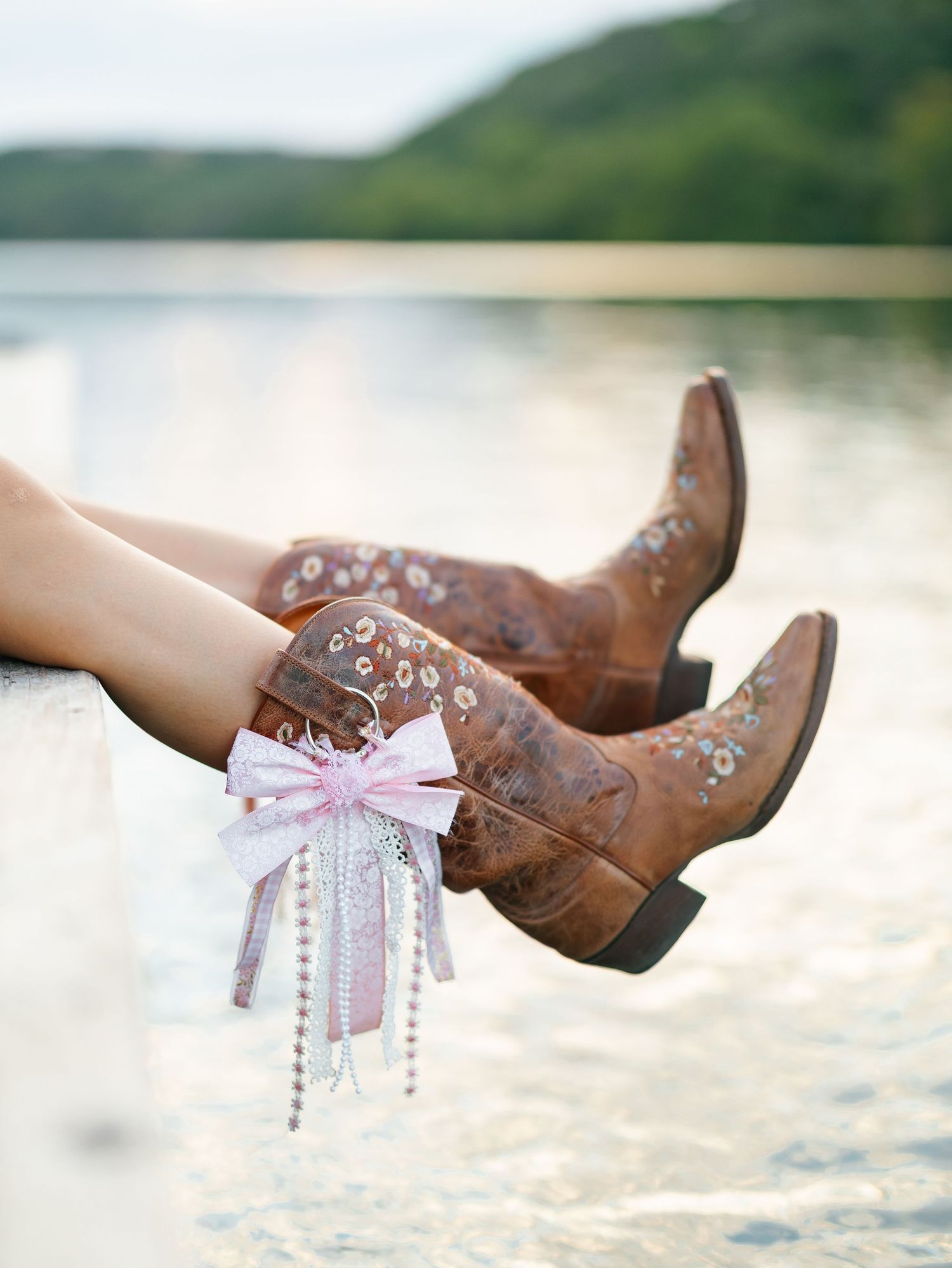 Brown cowboy boots with floral embroidery and pink bow ribbon detail, resting over a serene lake backdrop.