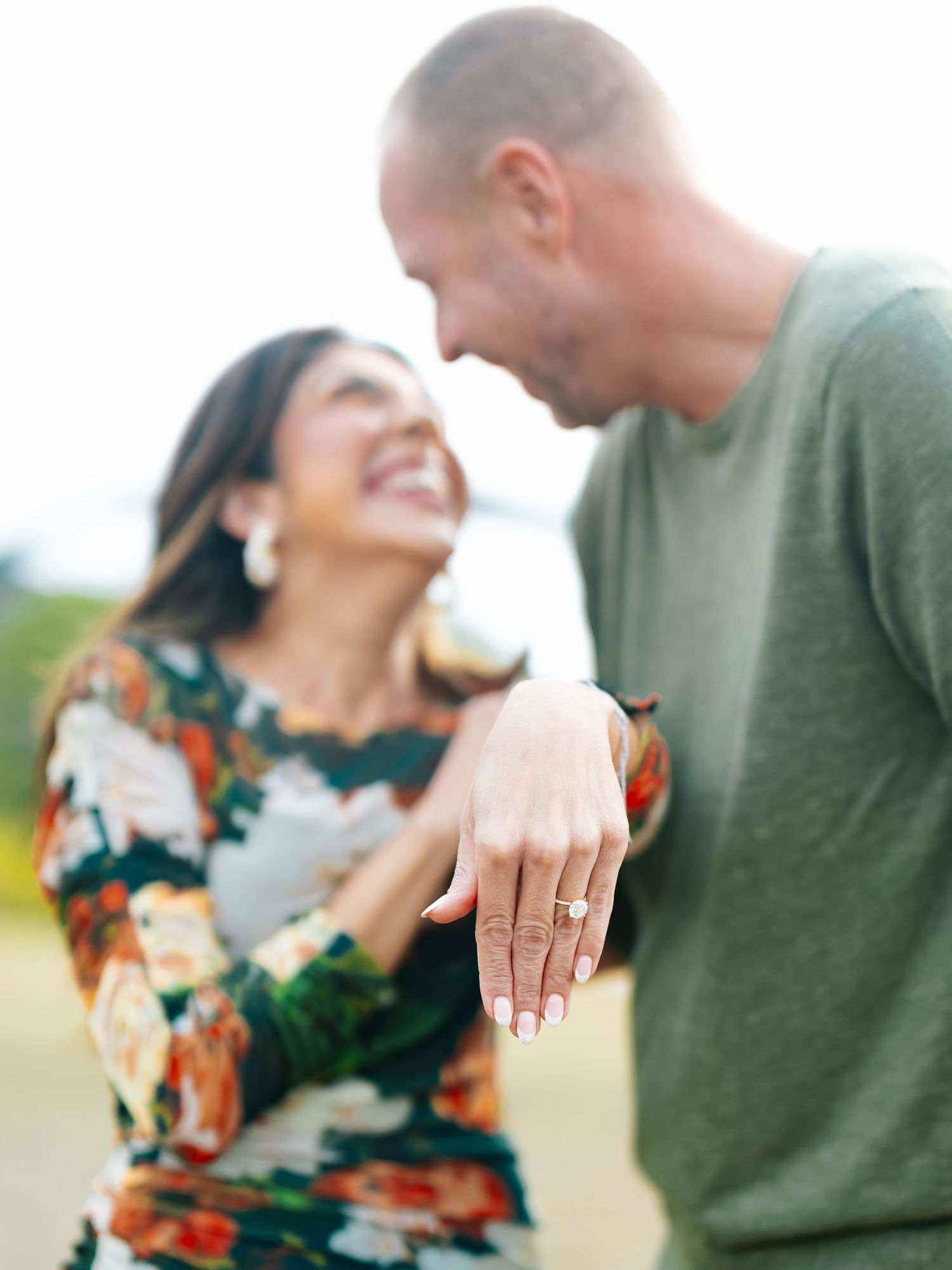 Joyful couple showing off an engagement ring with a blurred outdoor background.