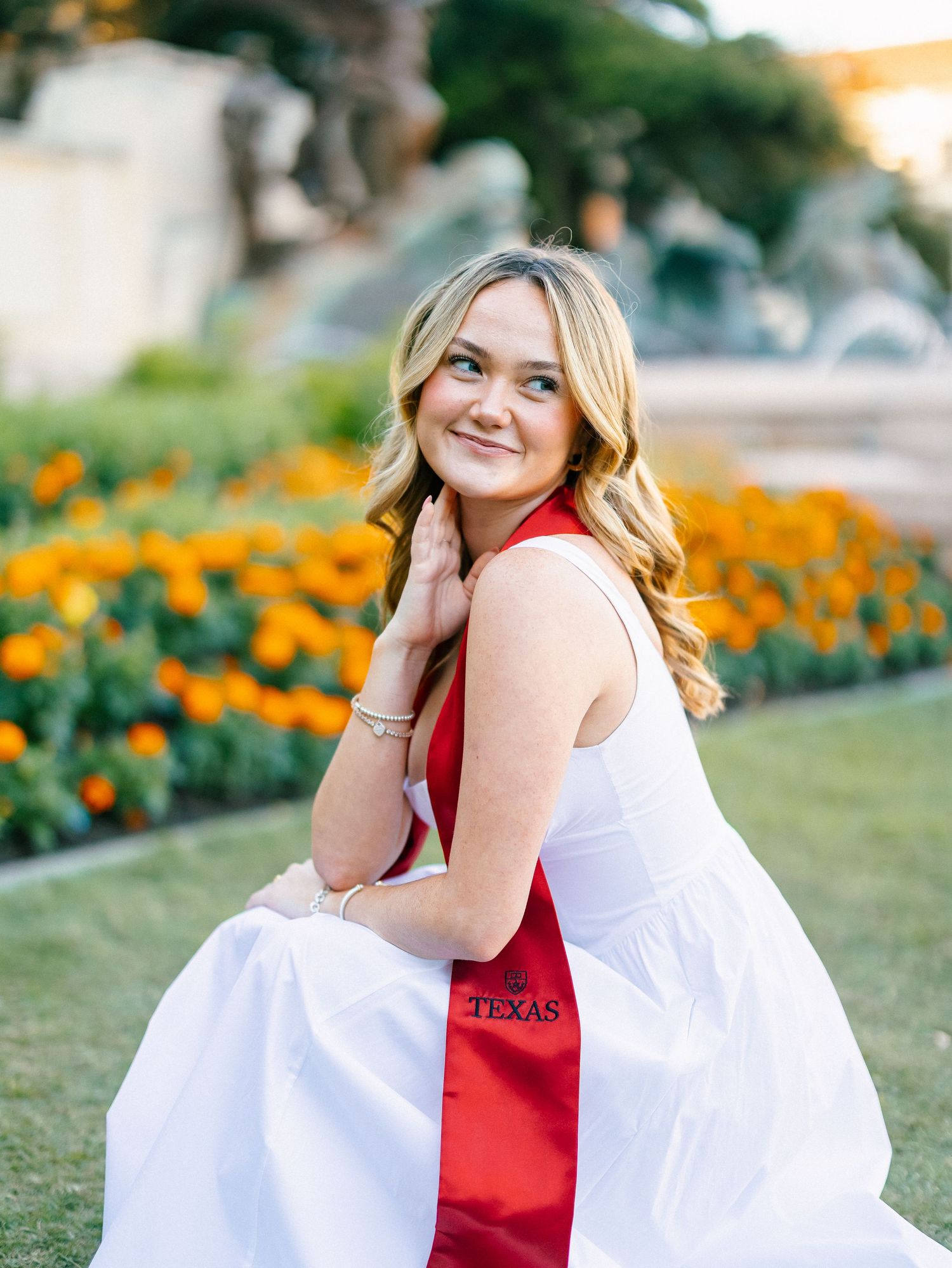 Young woman in white dress with red Texas sash smiles over shoulder, seated outdoors with orange flowers in background.