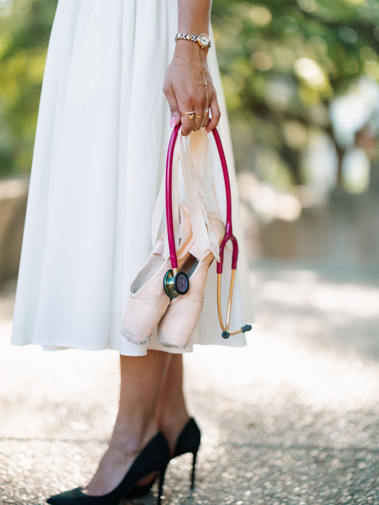 Woman in white dress holding pink ballet shoes and a magenta stethoscope, wearing black heels and gold jewelry.