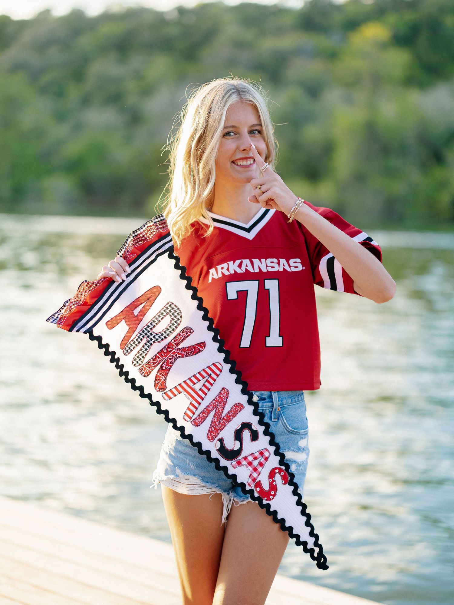 Blonde woman in Arkansas #71 red jersey holding a pennant flag on a lakeside dock in denim shorts.