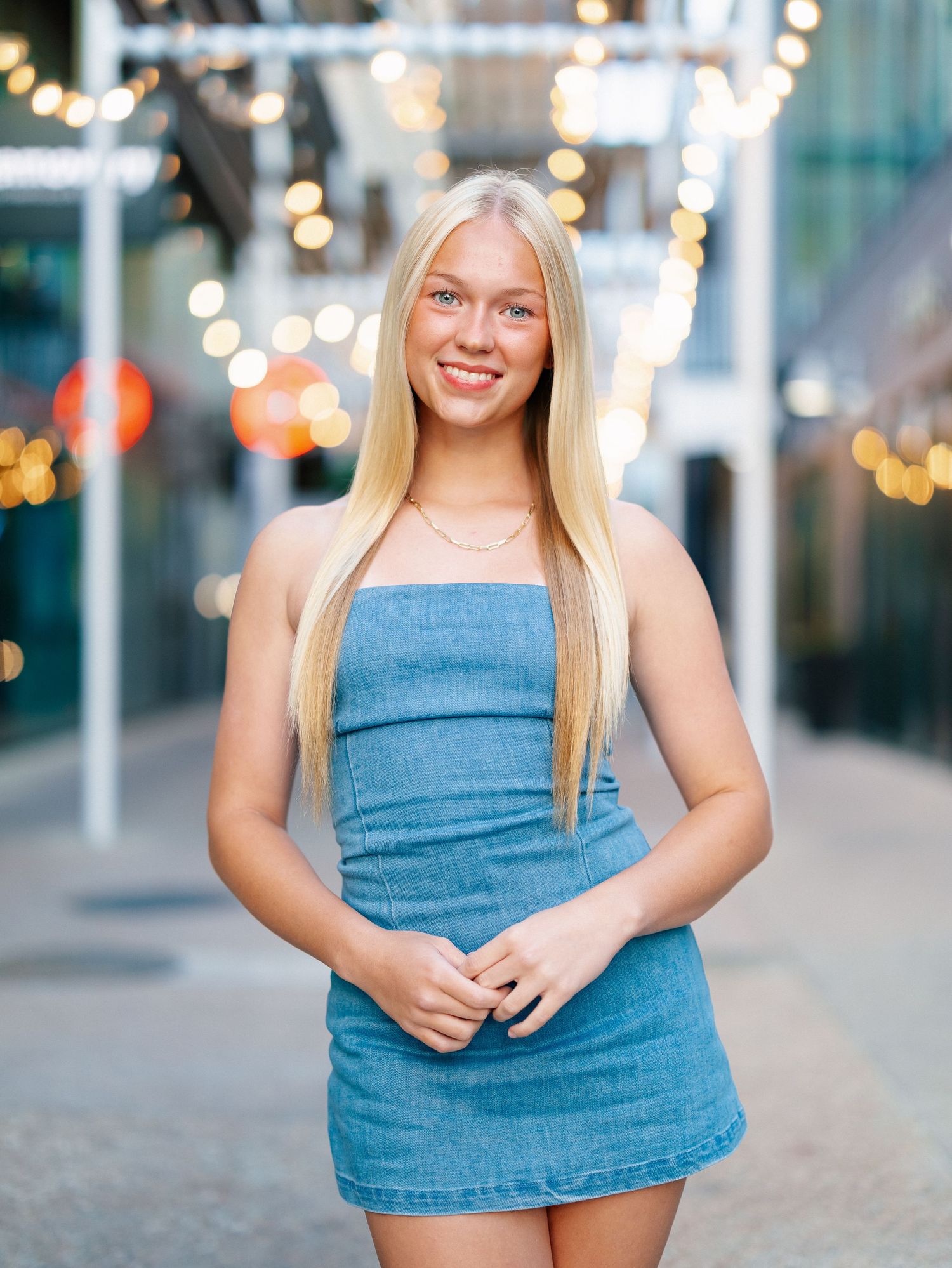 Young blonde woman smiling in a blue denim mini dress, posing outdoors with bokeh string lights in background.