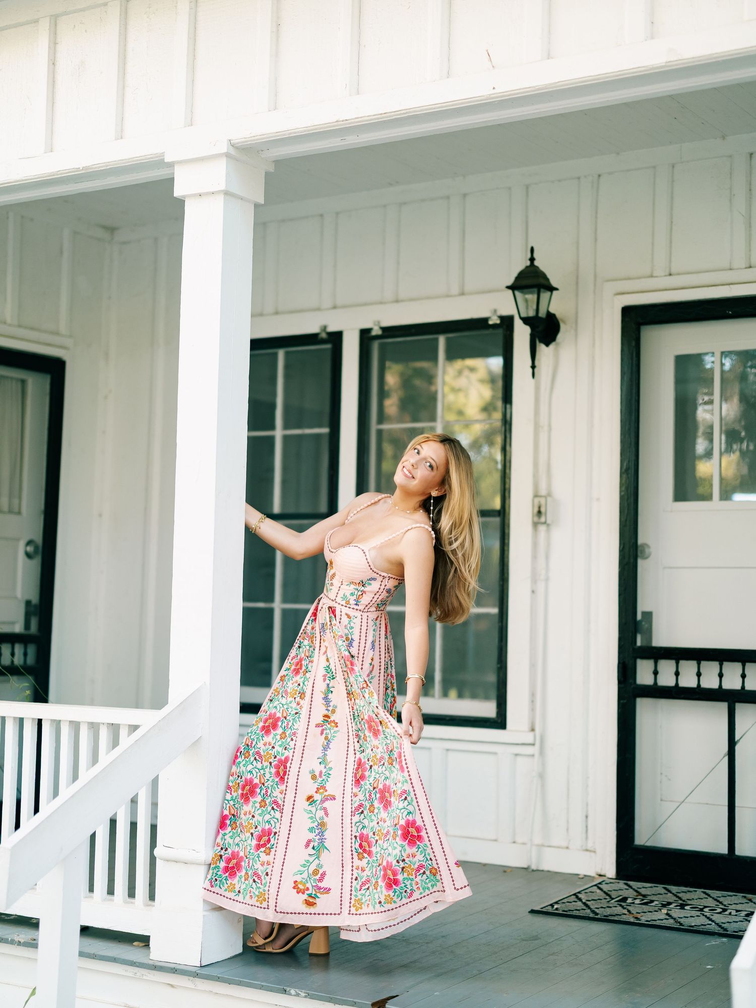 Blonde woman in pink floral maxi dress posing on white farmhouse porch steps.