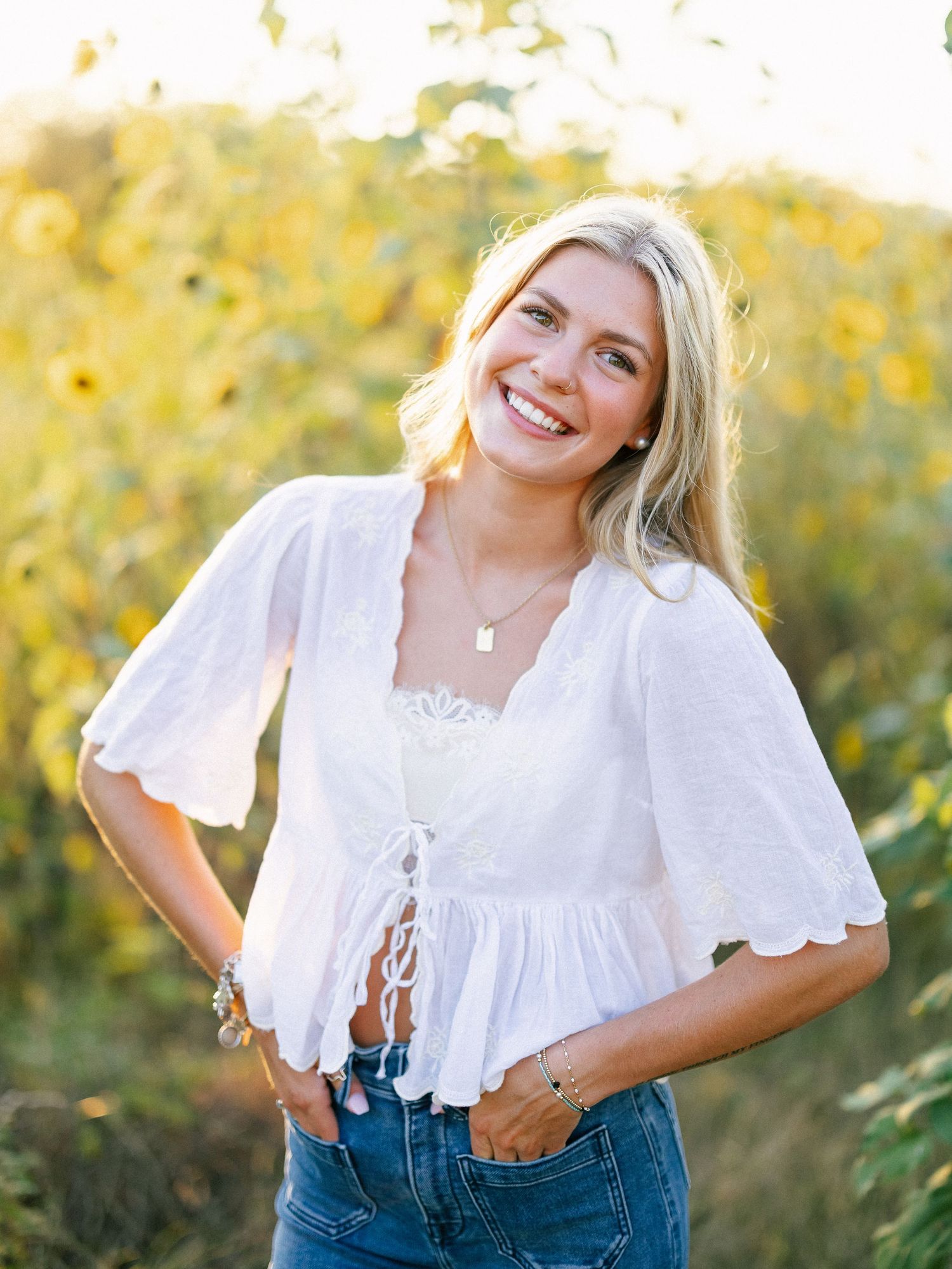 Smiling blonde woman in white lace top and jeans posing in a sunflower field on a sunny day.