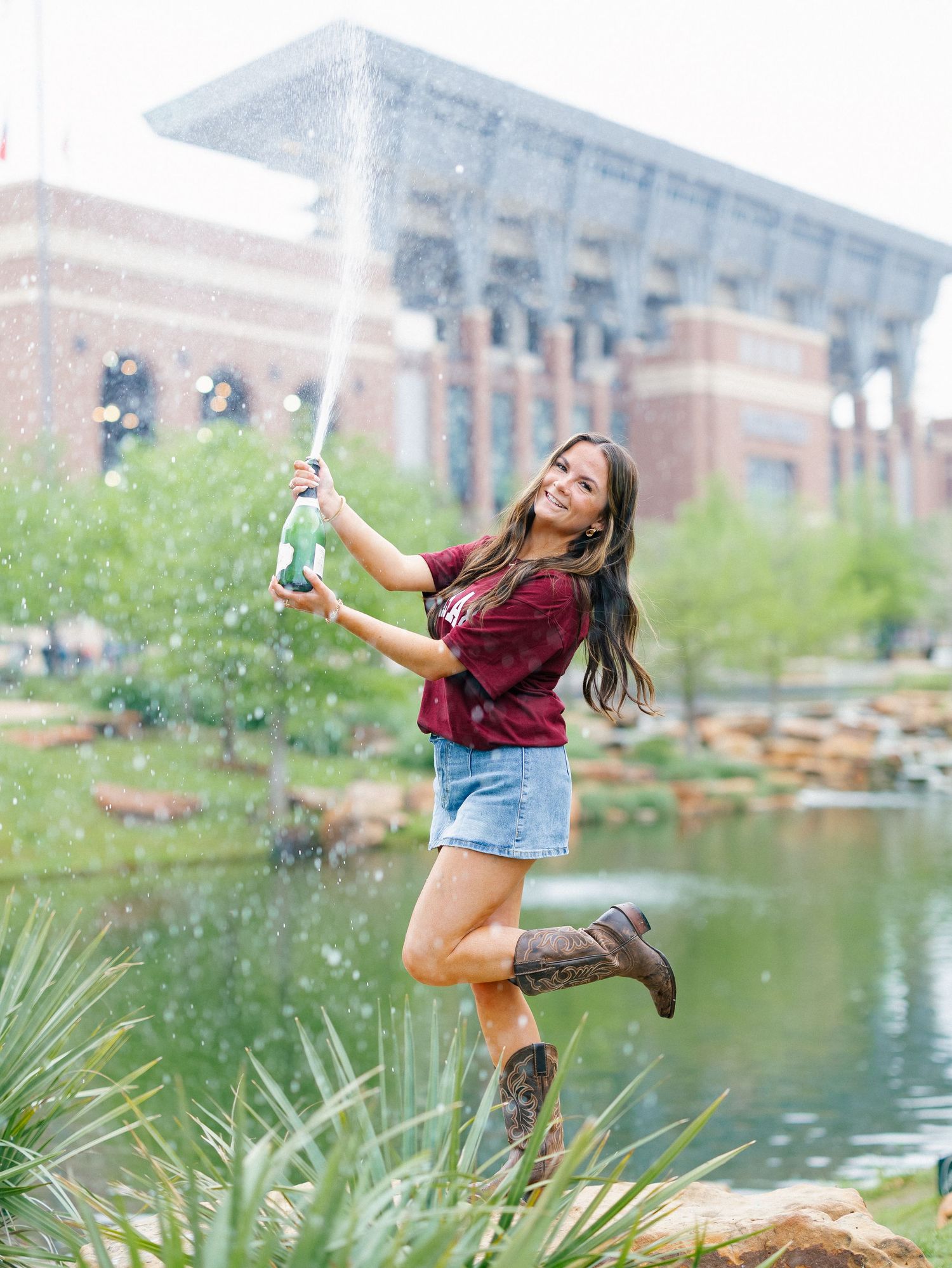 Young woman in maroon shirt and cowboy boots celebrates with champagne by a pond near a brick stadium.