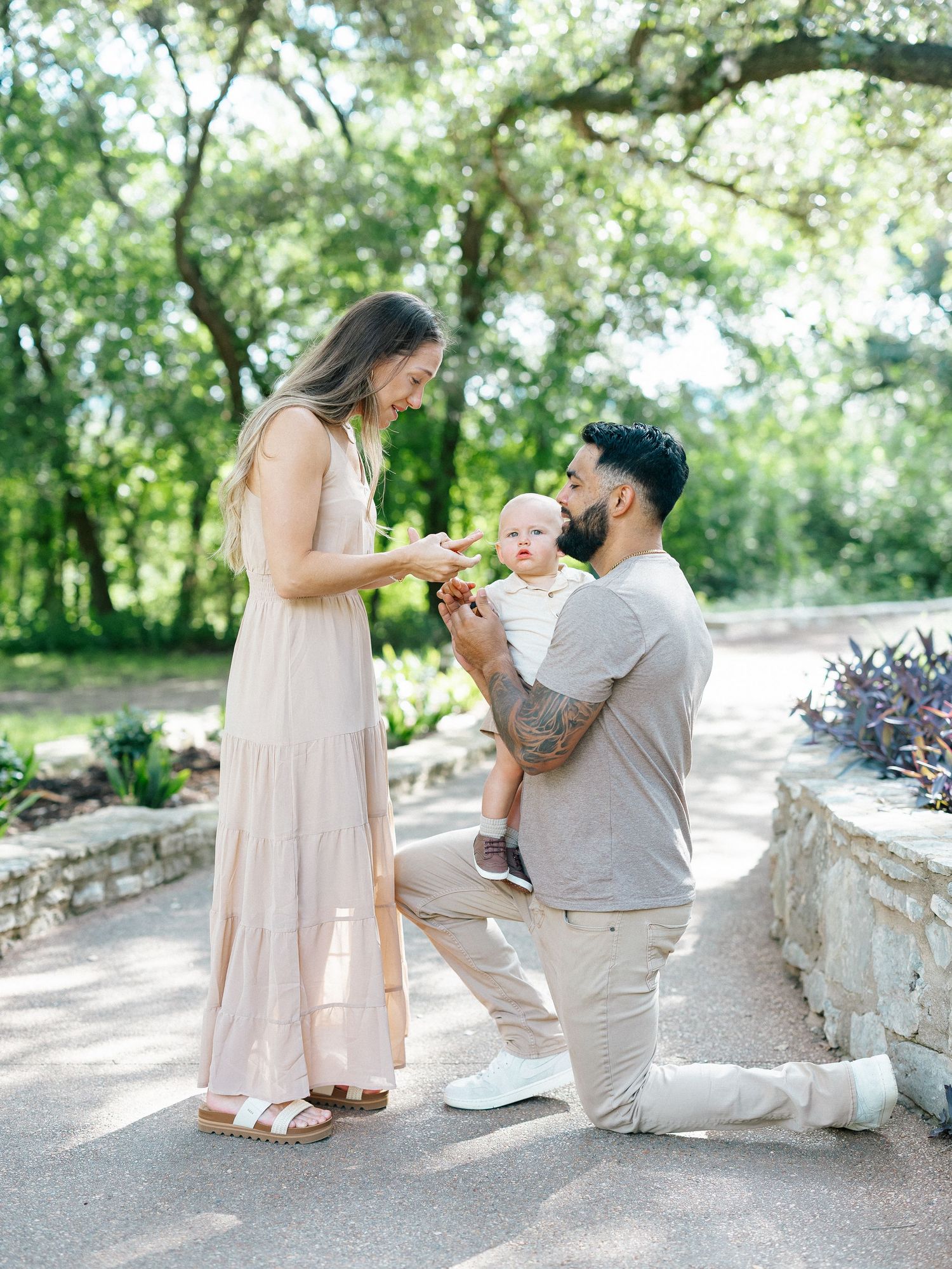 A man on one knee proposes to a woman in a blush dress while cradling their baby in a park.