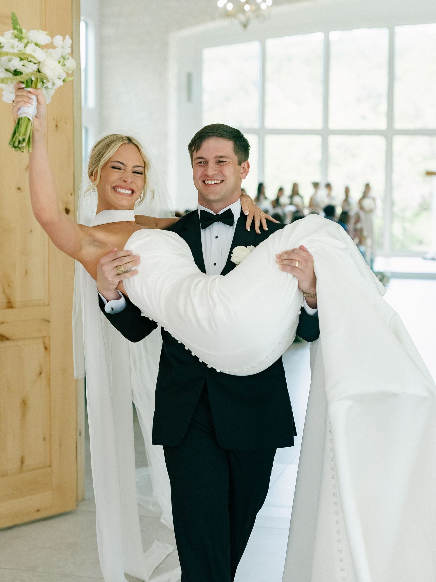 Joyful groom carrying bride in arms through barn wedding venue doorway.
