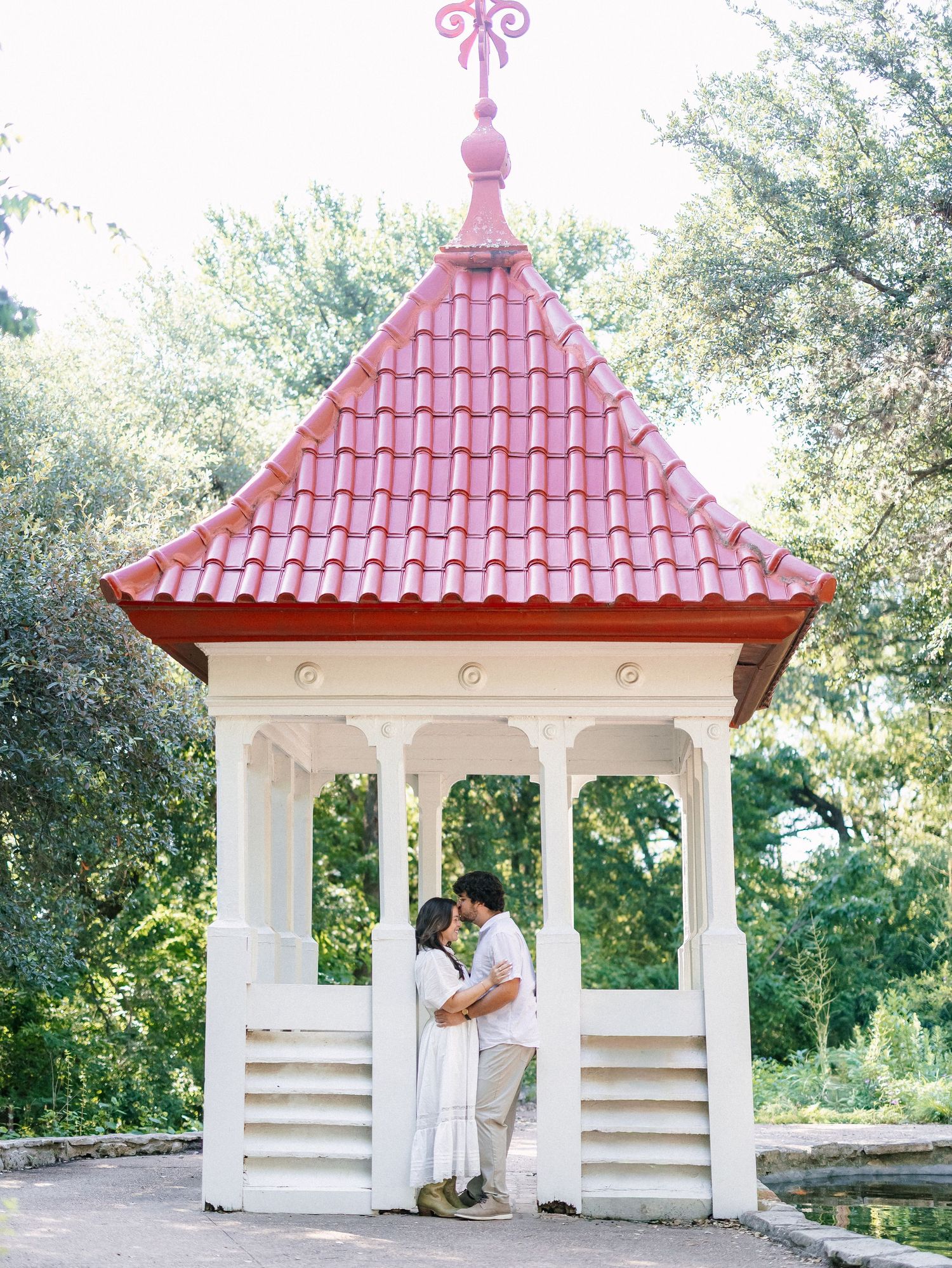 Couple shares a tender kiss inside a charming white gazebo with a red tiled roof surrounded by trees.
