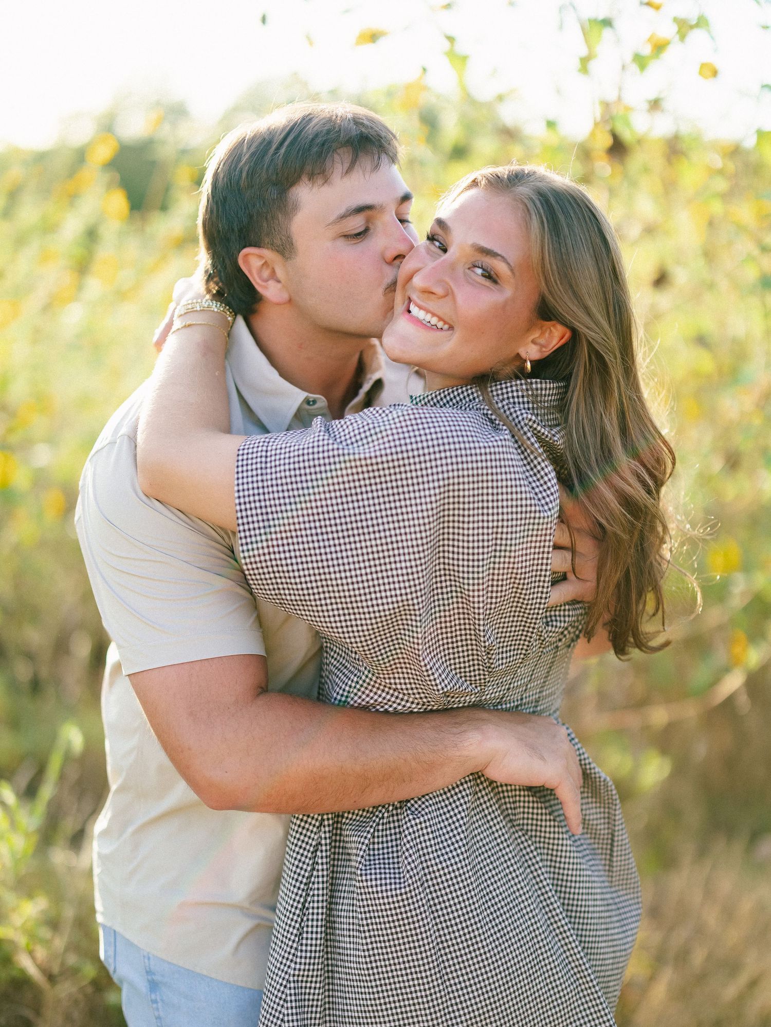 Man kissing woman on the cheek while embracing in a sunny wildflower field during engagement photo session.