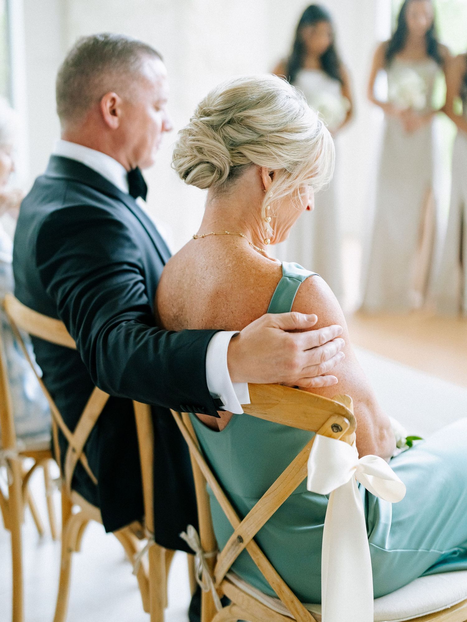 Groom in black tuxedo tenderly places hand on shoulder of woman in sage green dress during wedding ceremony.