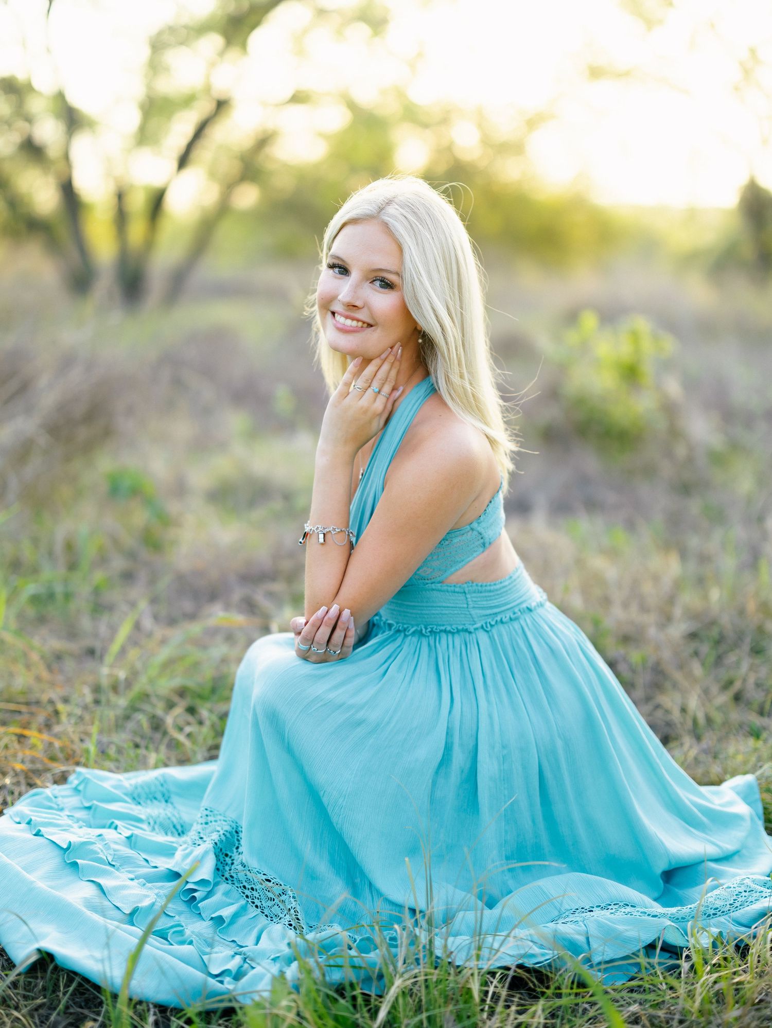 Blonde woman in a flowing turquoise halter dress sitting gracefully in a sunlit grassy field.