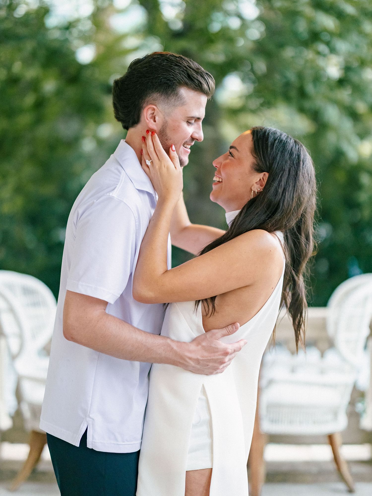 Happy engaged couple embracing and smiling at each other outdoors with lush green trees in background.