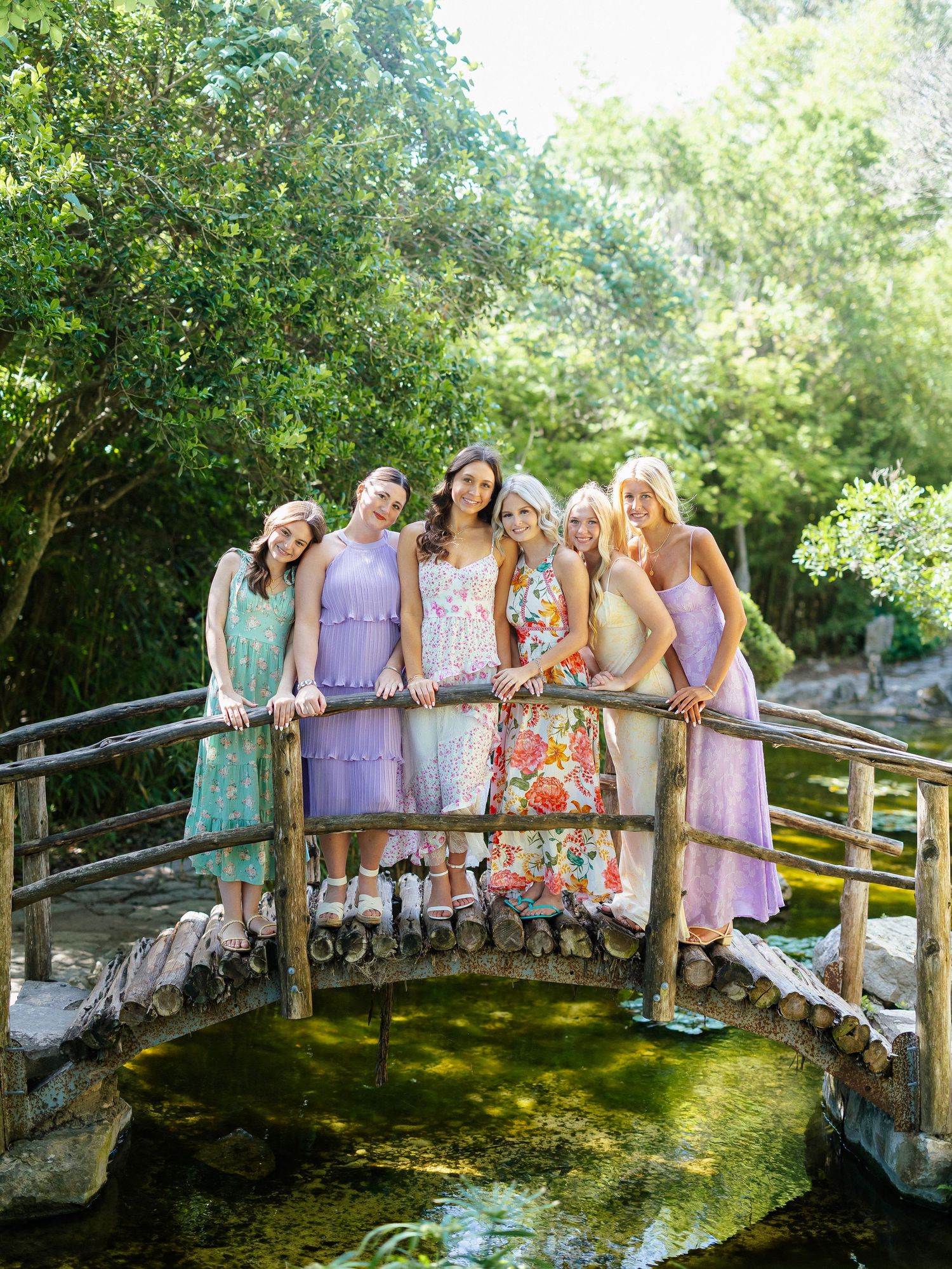 Five bridesmaids in pastel purple and floral dresses pose together on a rustic wooden garden bridge.