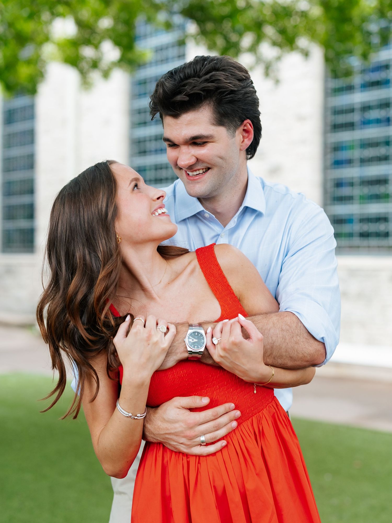 Newly engaged couple embracing and smiling at each other outdoors, woman wearing red dress showing engagement ring.