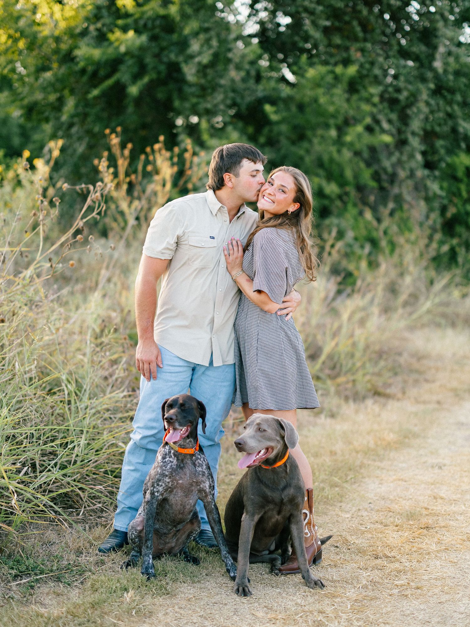 Couple sharing a kiss outdoors on a dirt path with two brown dogs sitting in front of them.