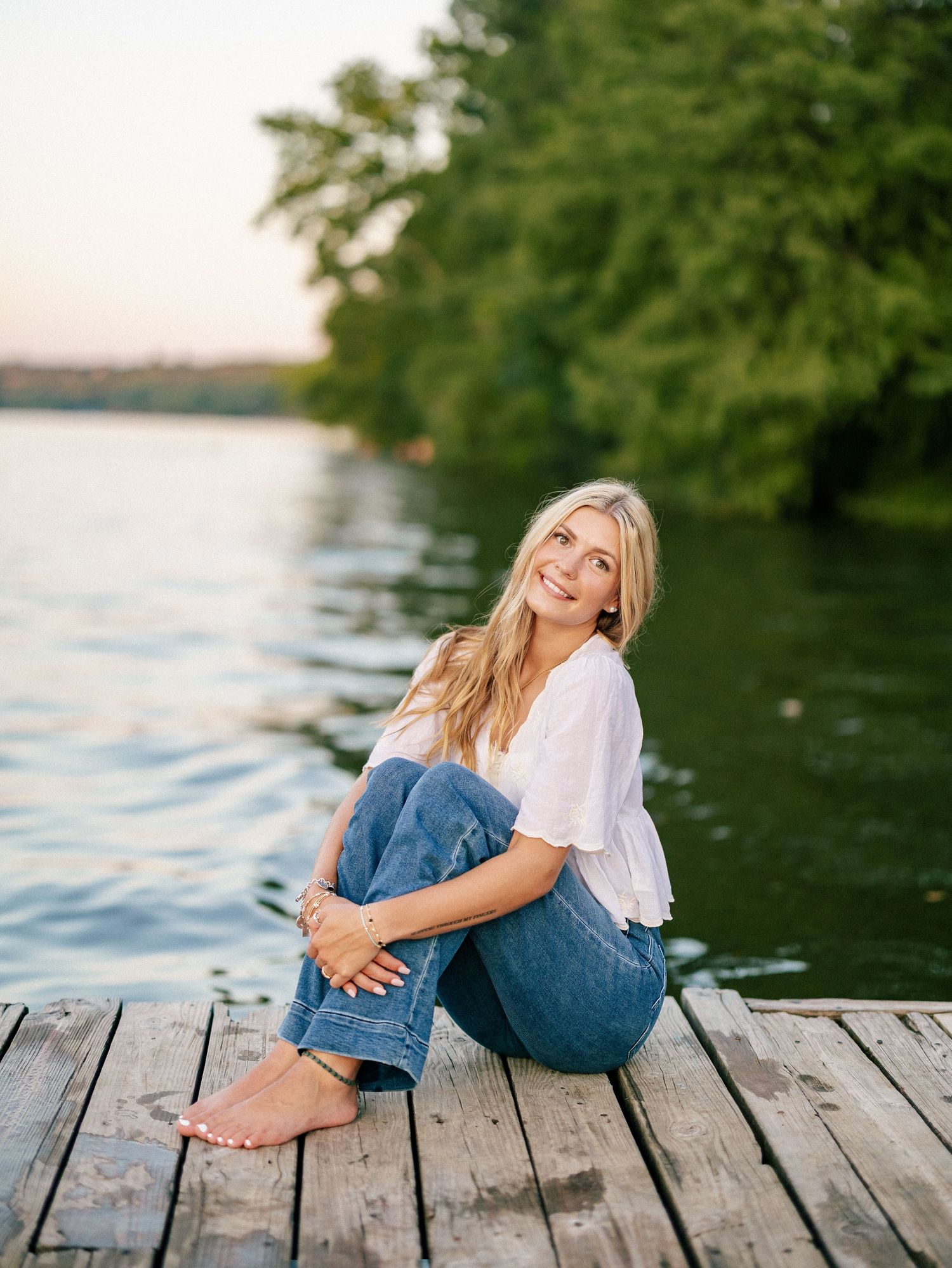 Smiling blonde woman sitting barefoot on a wooden dock by a calm lake, wearing jeans and a white top.