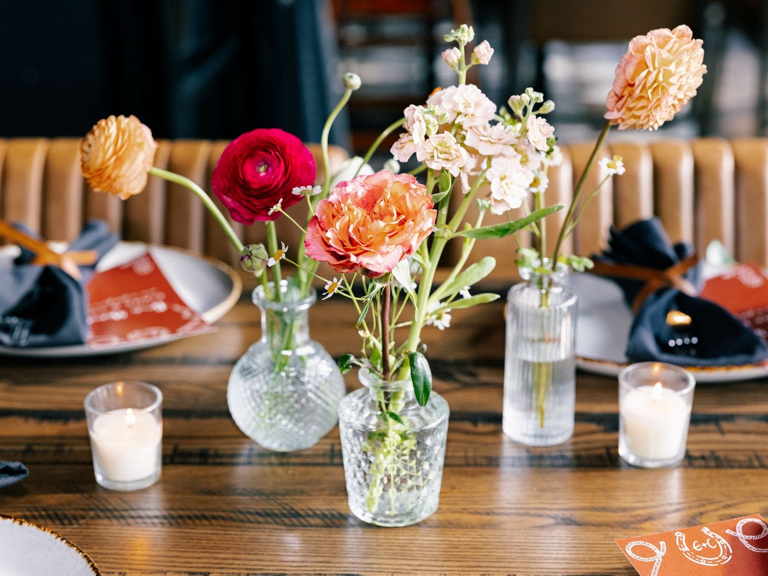 Colorful floral centerpiece with ranunculus and dahlias in glass bud vases on a wooden dining table.