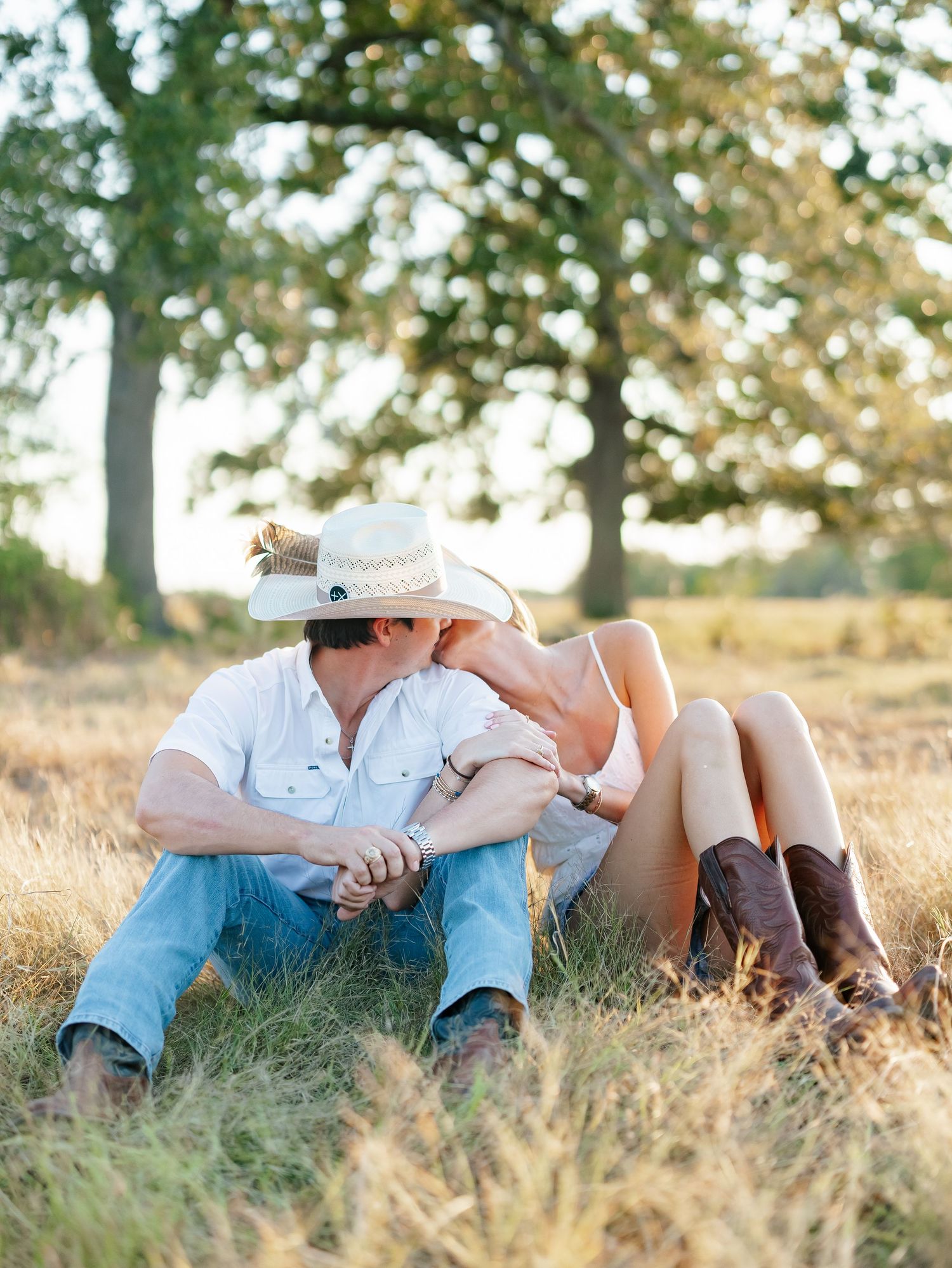 A couple shares a kiss while sitting in a golden meadow beneath oak trees at sunset.