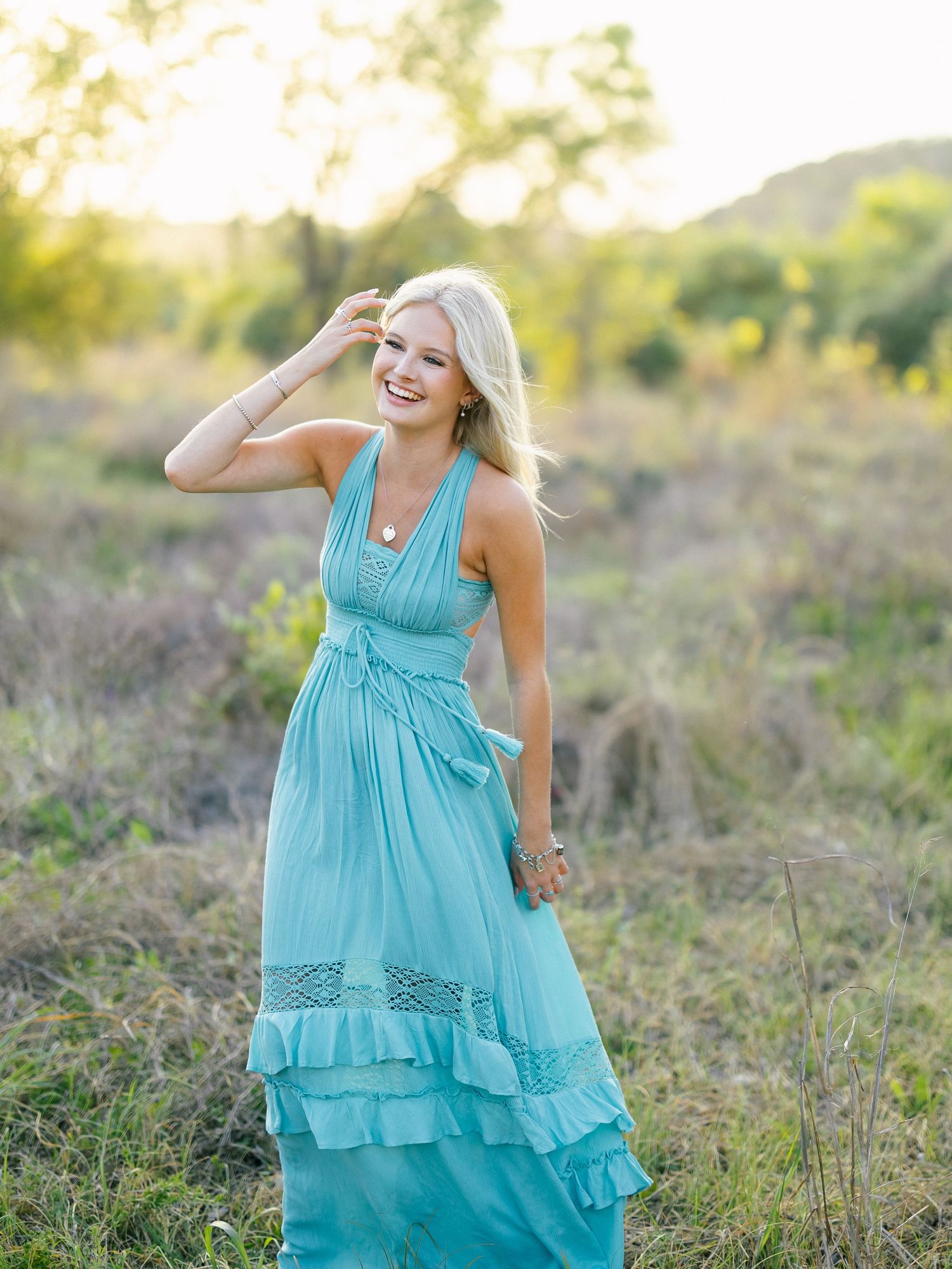 Smiling blonde woman in a teal maxi dress standing in a sunlit field during golden hour.
