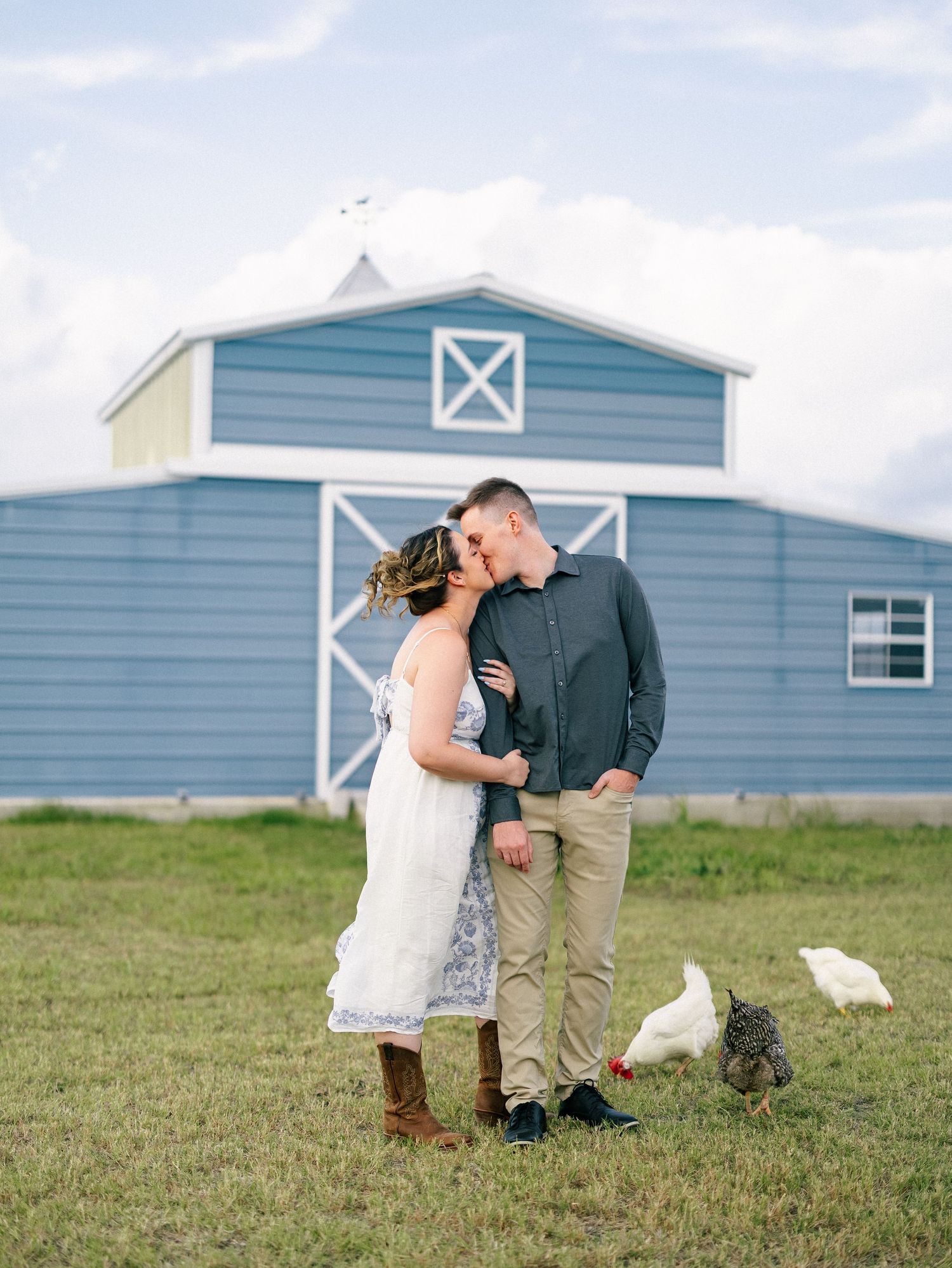 Couple kissing in front of a blue barn with white trim on a farm, surrounded by chickens and green grass.