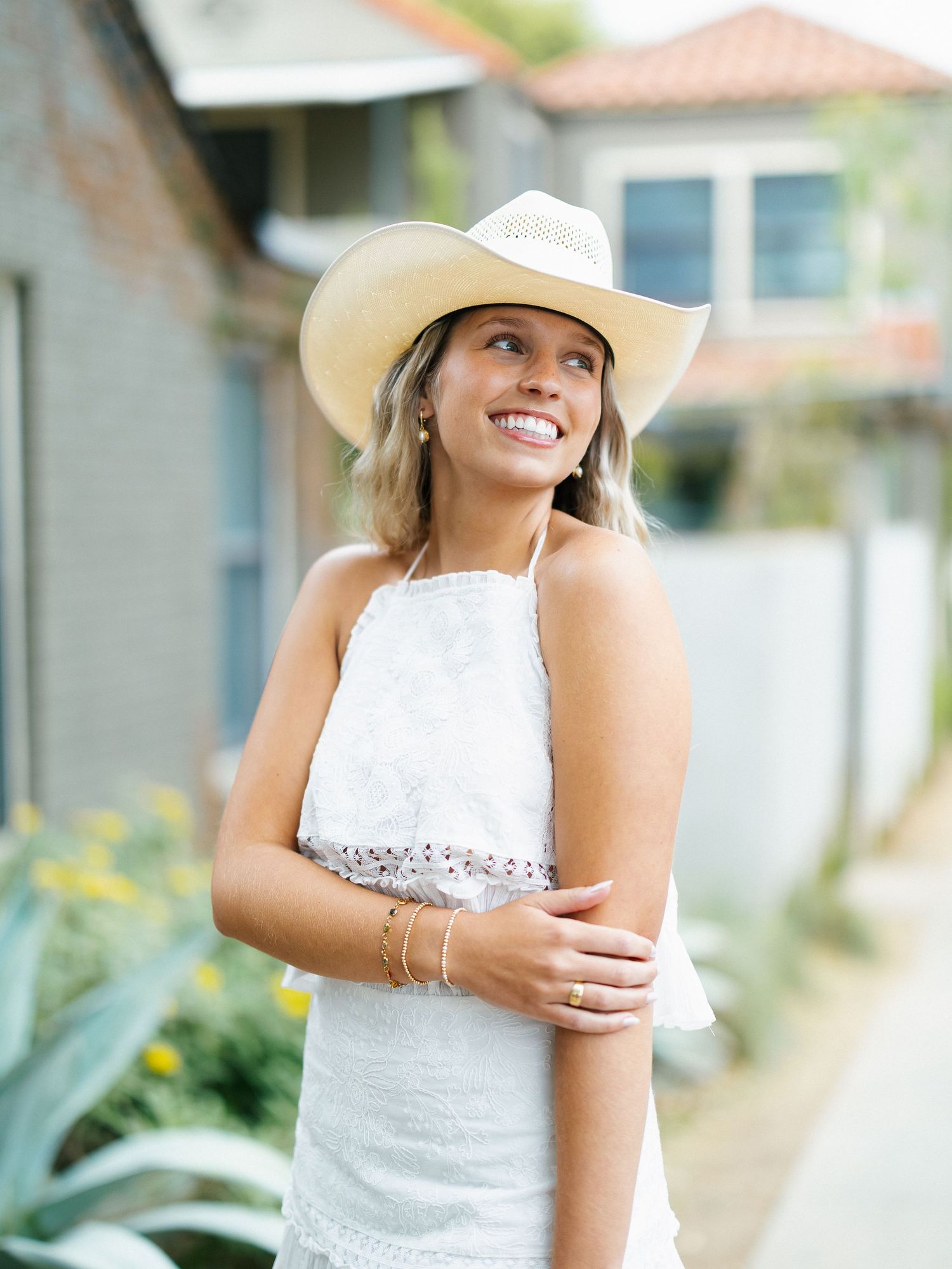 Smiling woman wearing a white cowboy hat and white lace halter top, posing outdoors near a house.