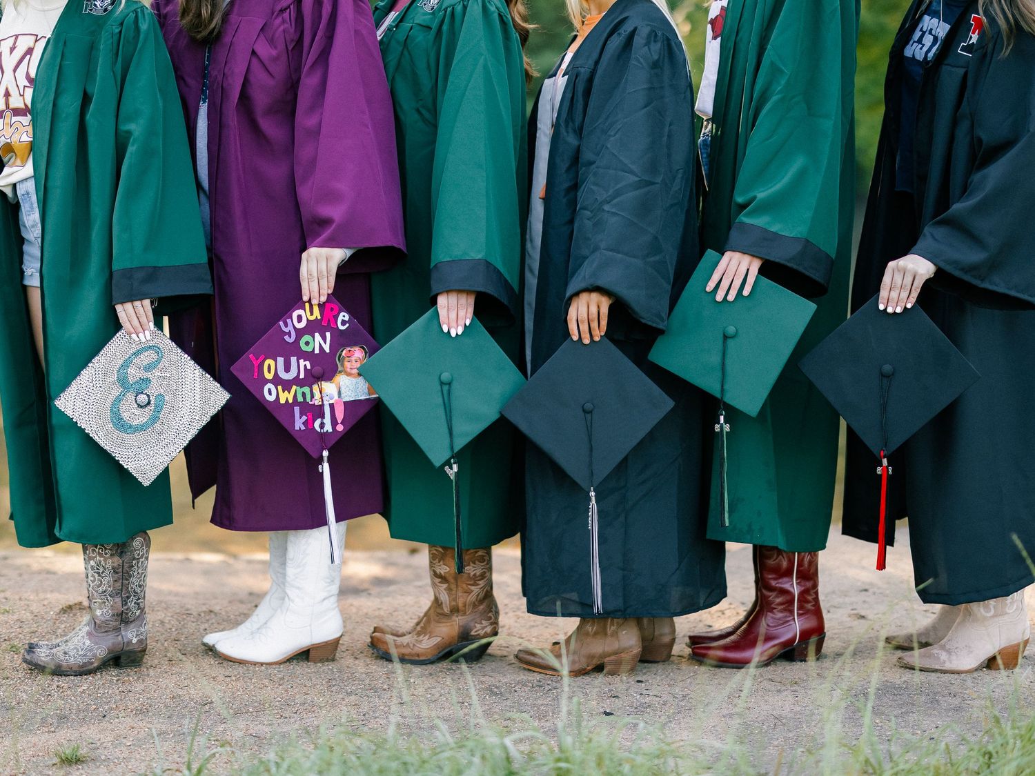Graduates in green and purple robes holding decorated caps, wearing cowboy boots at an outdoor graduation ceremony.