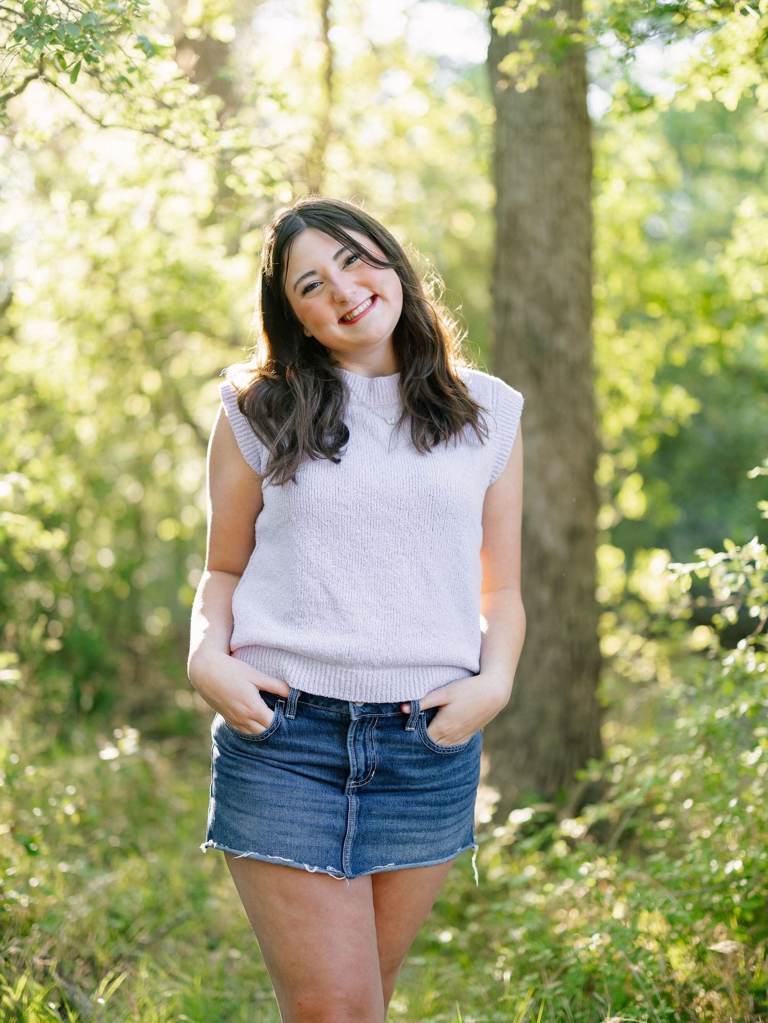 Smiling young woman in white sleeveless top and denim skirt posing in green forest.