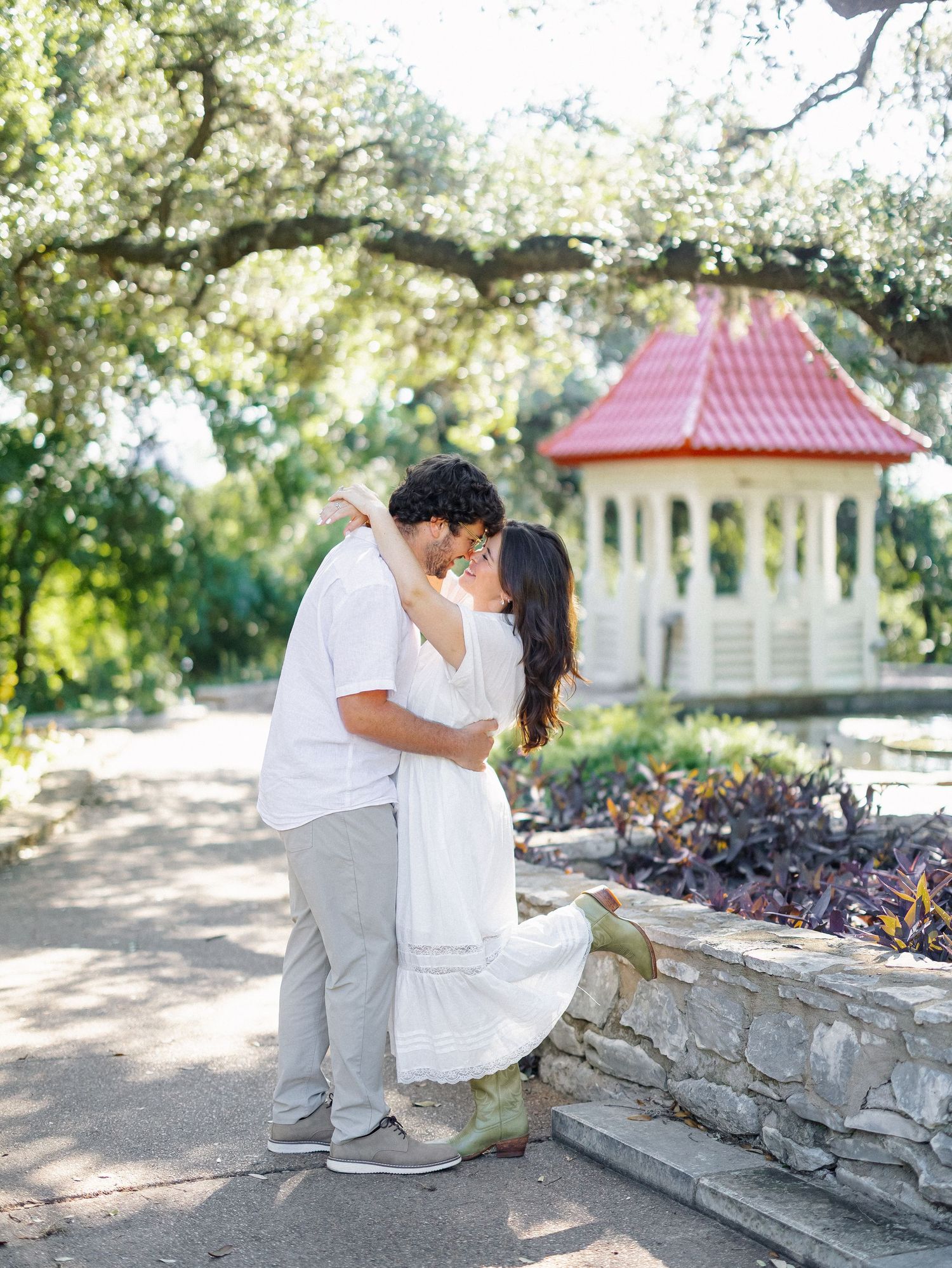 Couple sharing a romantic kiss in a sunlit garden with a red-roofed gazebo and lush greenery in the background.