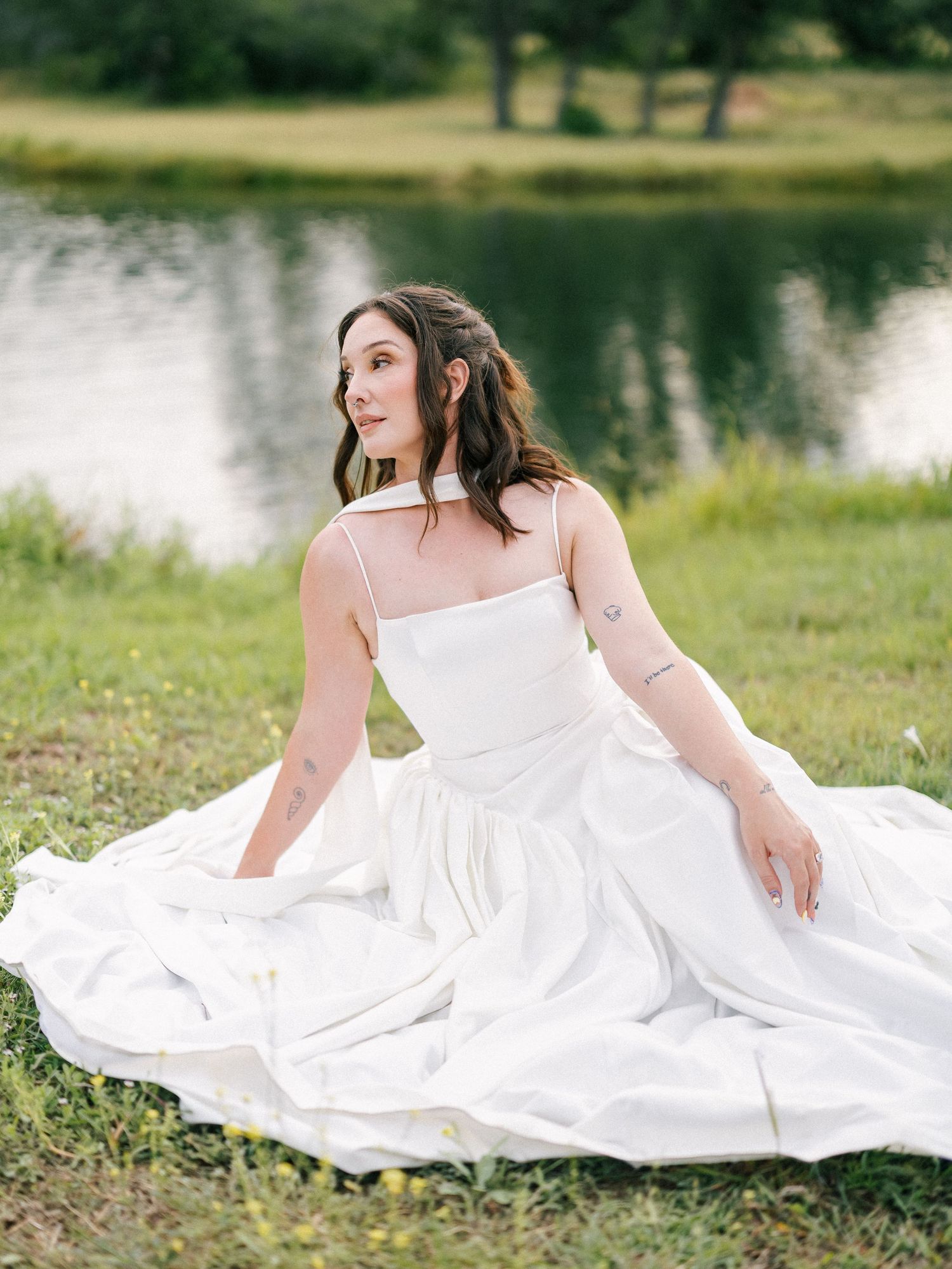 Bride in a flowing white gown sits gracefully on green grass beside a serene pond, gazing softly to the side.