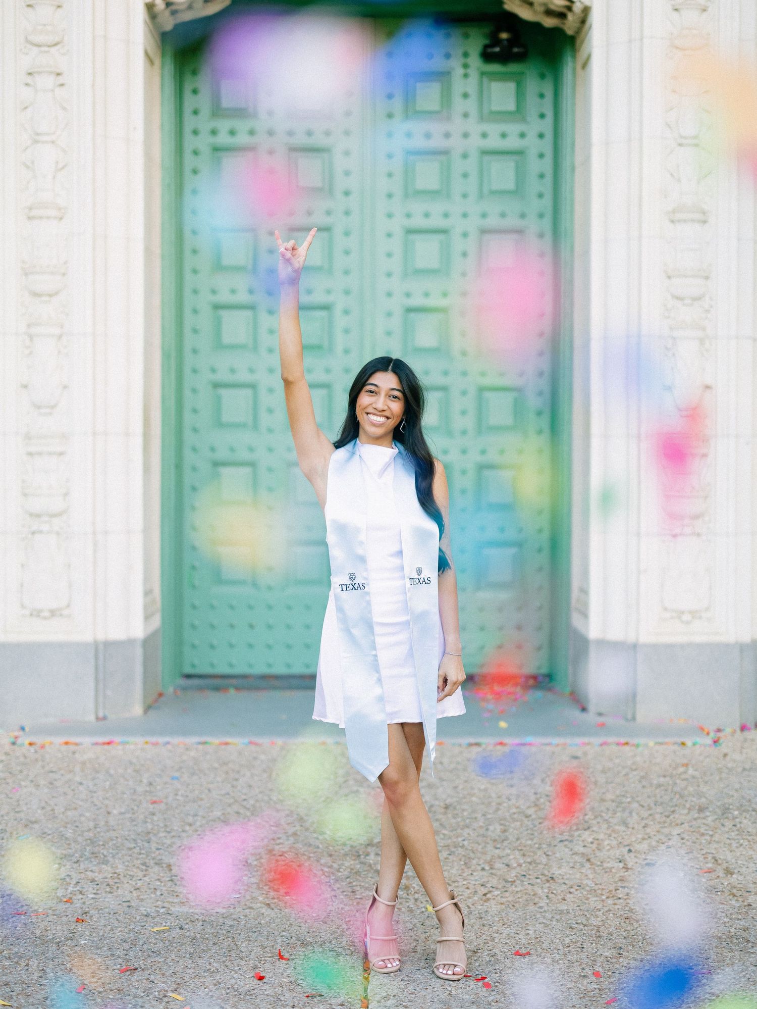 Young woman in white dress celebrating with colorful confetti in front of a green ornate door.
