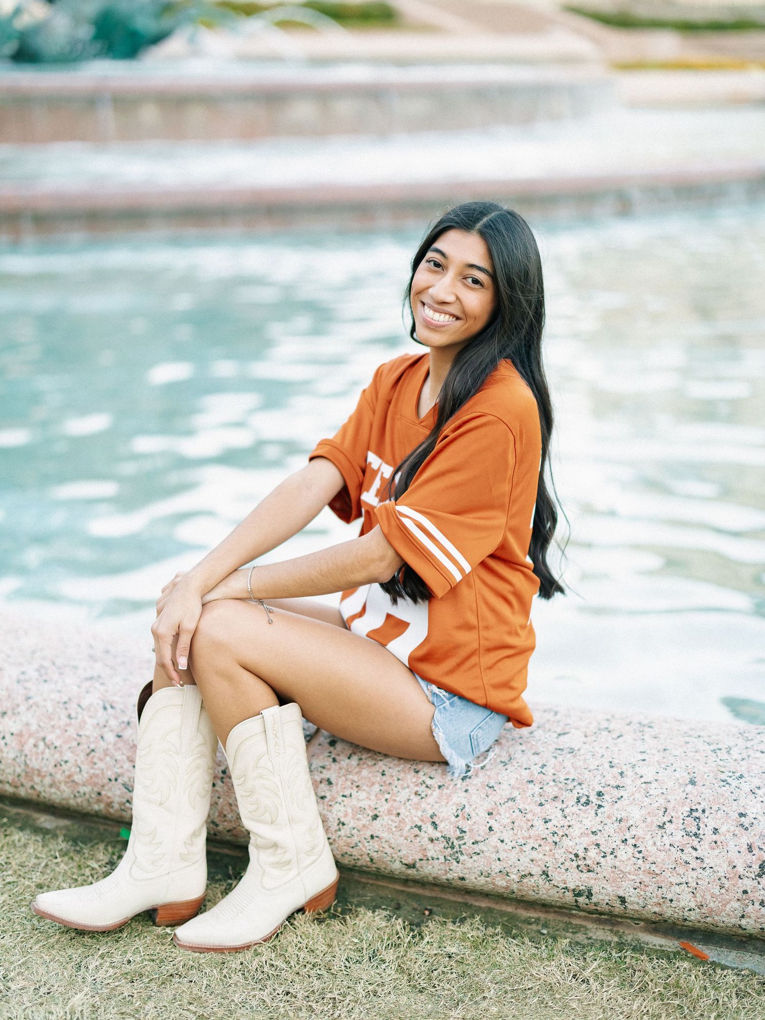 Young woman in orange jersey and white cowboy boots sitting by a fountain, smiling outdoors.