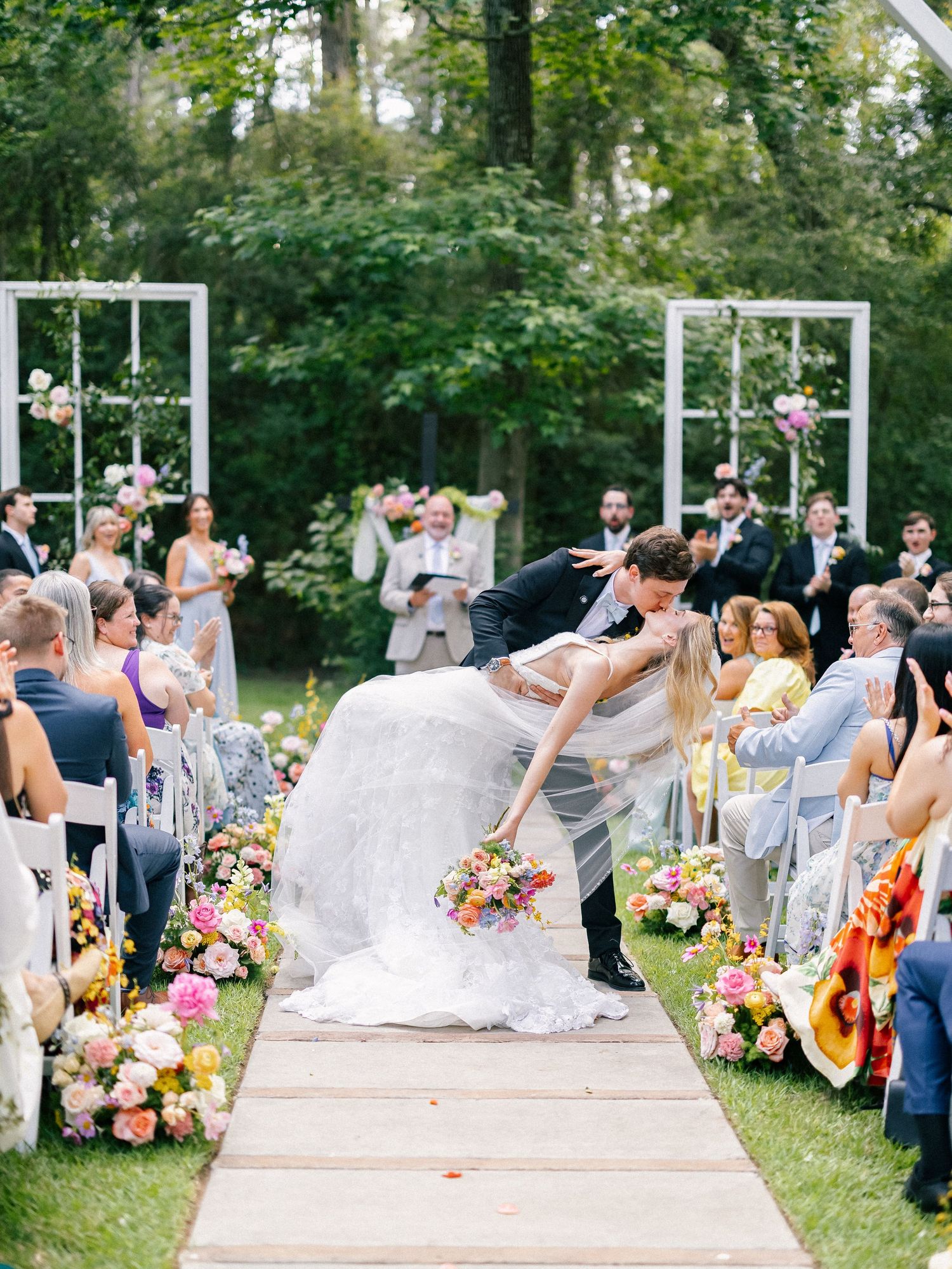 Bride and groom share a romantic dip kiss at outdoor wedding ceremony aisle lined with colorful floral arrangements.
