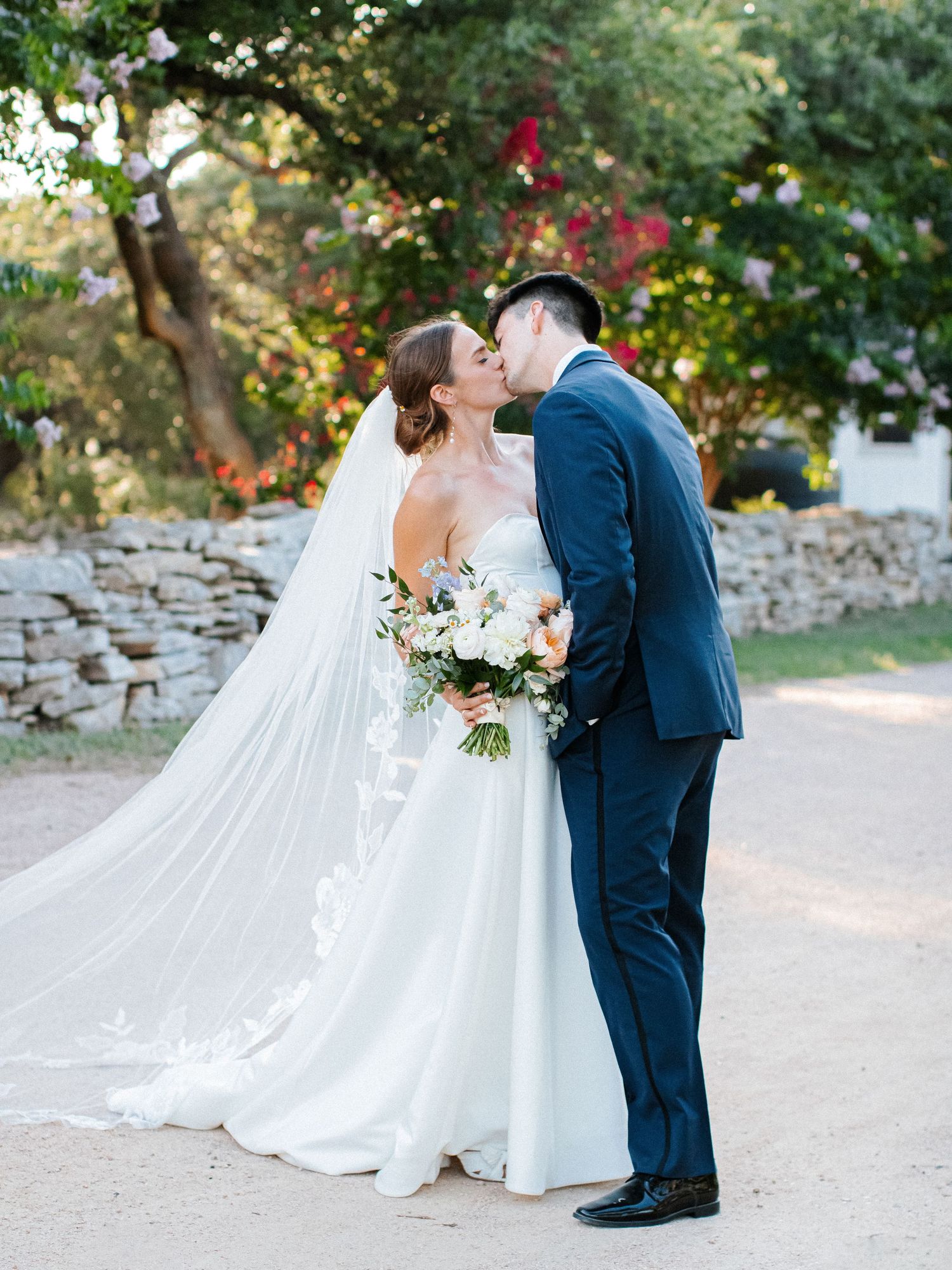 Newlyweds kiss on gravel path surrounded by lush trees and stone wall at outdoor venue.