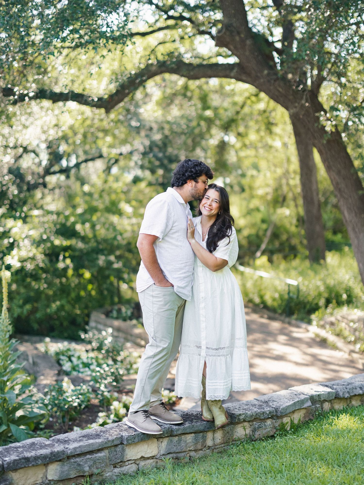 Couple shares a romantic moment standing on a stone wall surrounded by lush greenery, woman in white dress smiling.