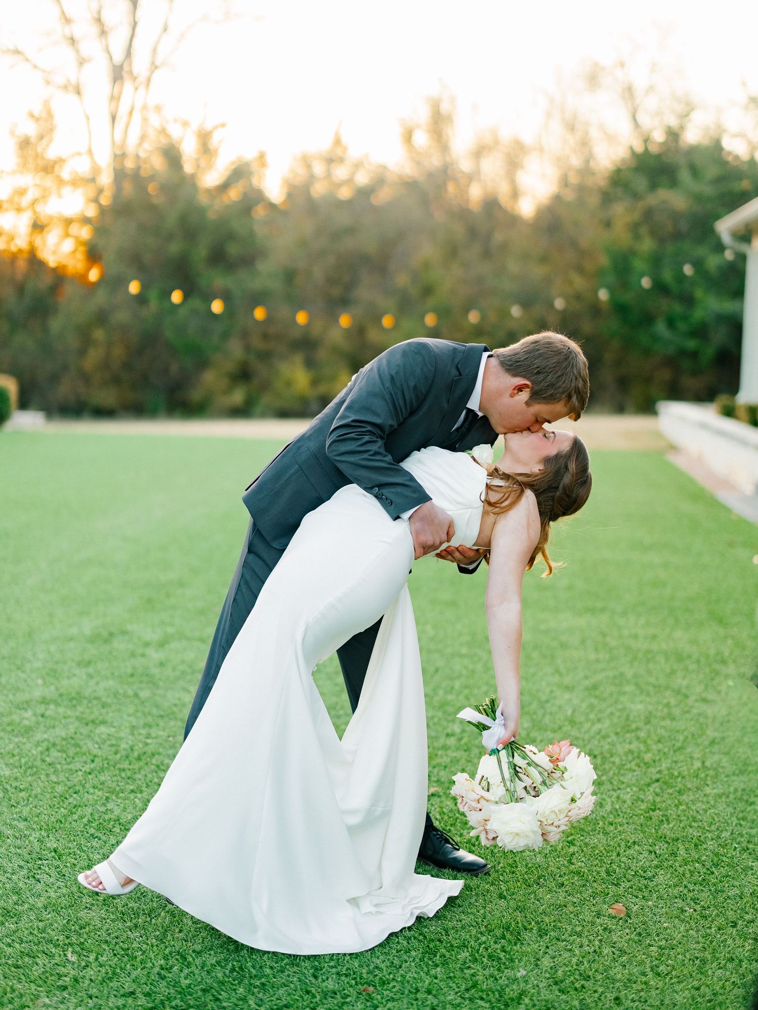 Groom dips bride for a romantic kiss on a green lawn at golden hour during their outdoor wedding.
