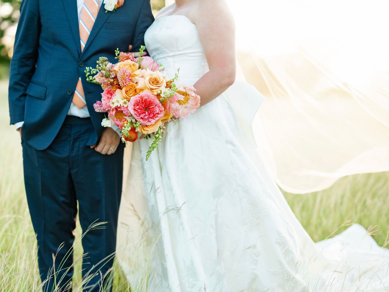 Bride and groom embracing in sunlit field, bride's veil flowing with colorful bridal bouquet.