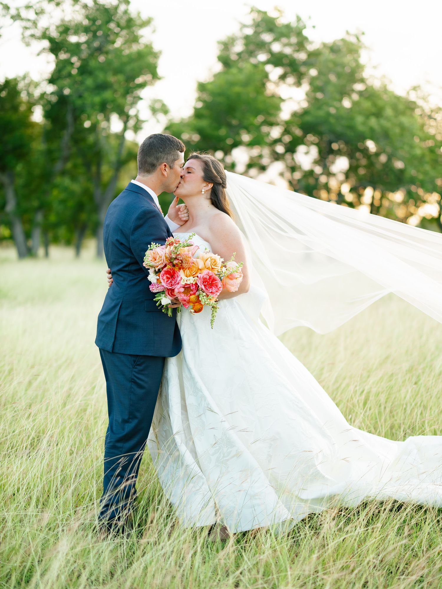 Bride and groom sharing a romantic kiss in a green meadow, her veil flowing in the breeze with a colorful bouquet.