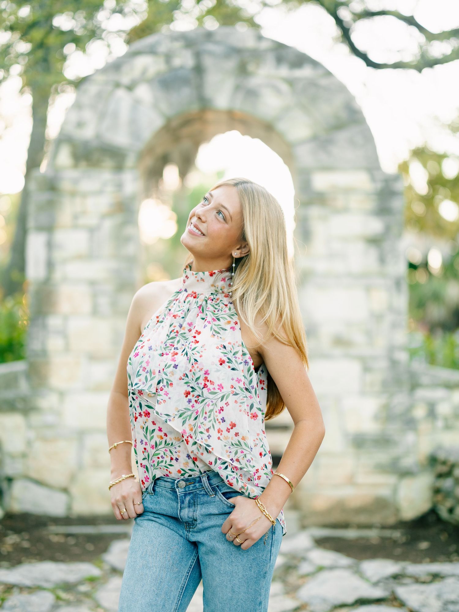 Smiling blonde woman in floral halter top and jeans before stone arch garden wall.