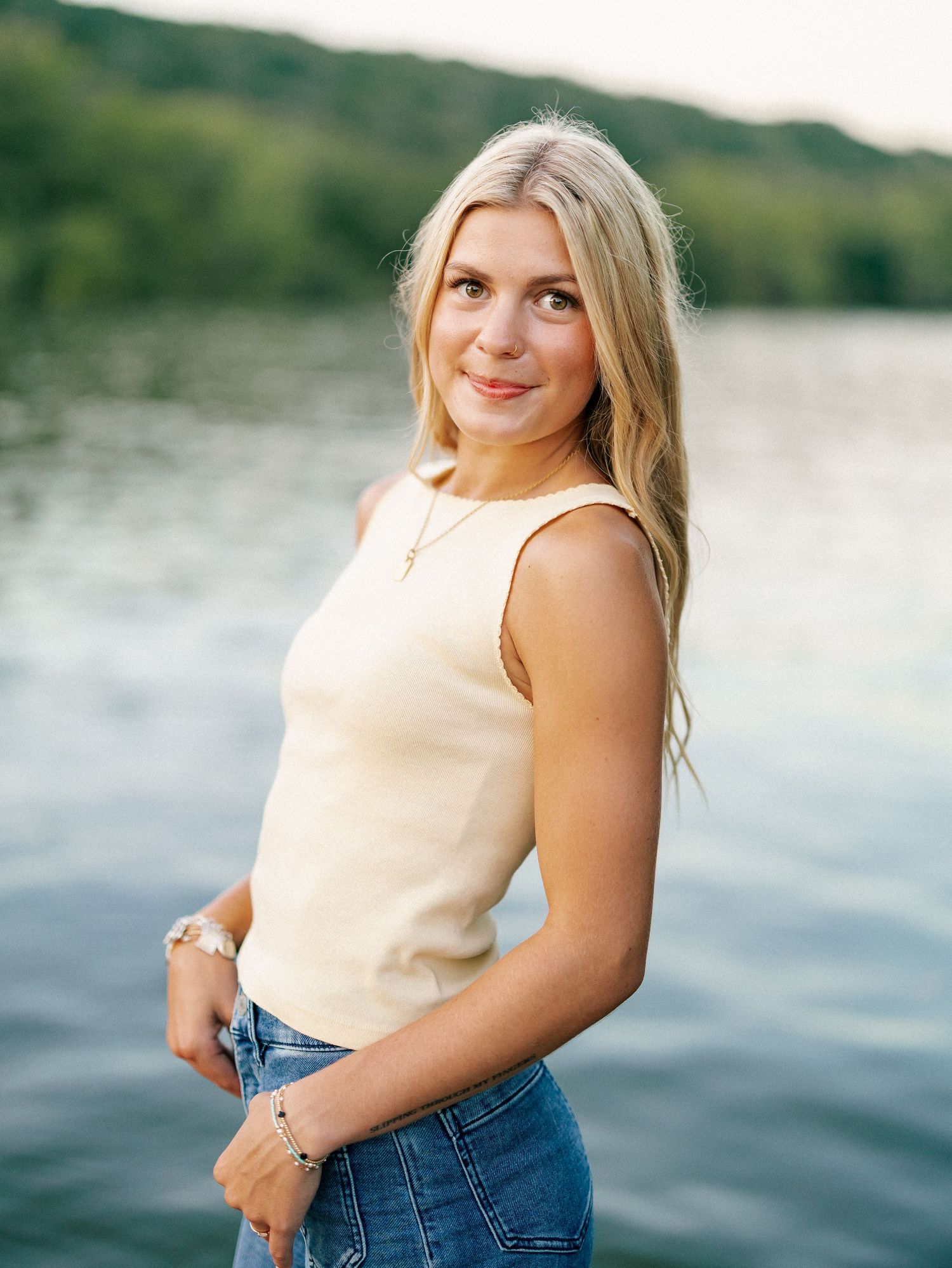 Young blonde woman in cream tank top and jeans posing by a lake with lush green hills in background.