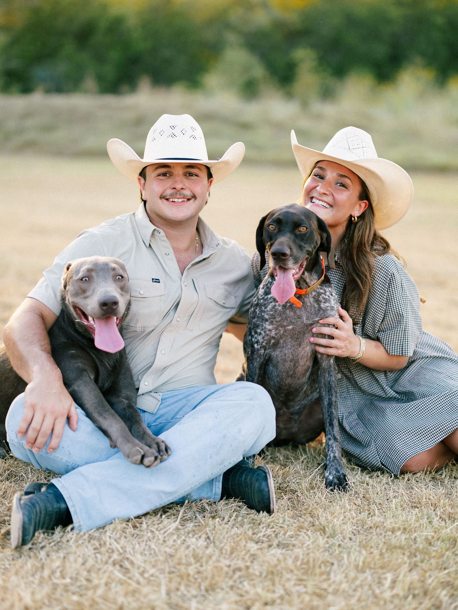 Smiling couple in cowboy hats sitting on grass with two dogs, a gray pit bull and a spotted pointer.
