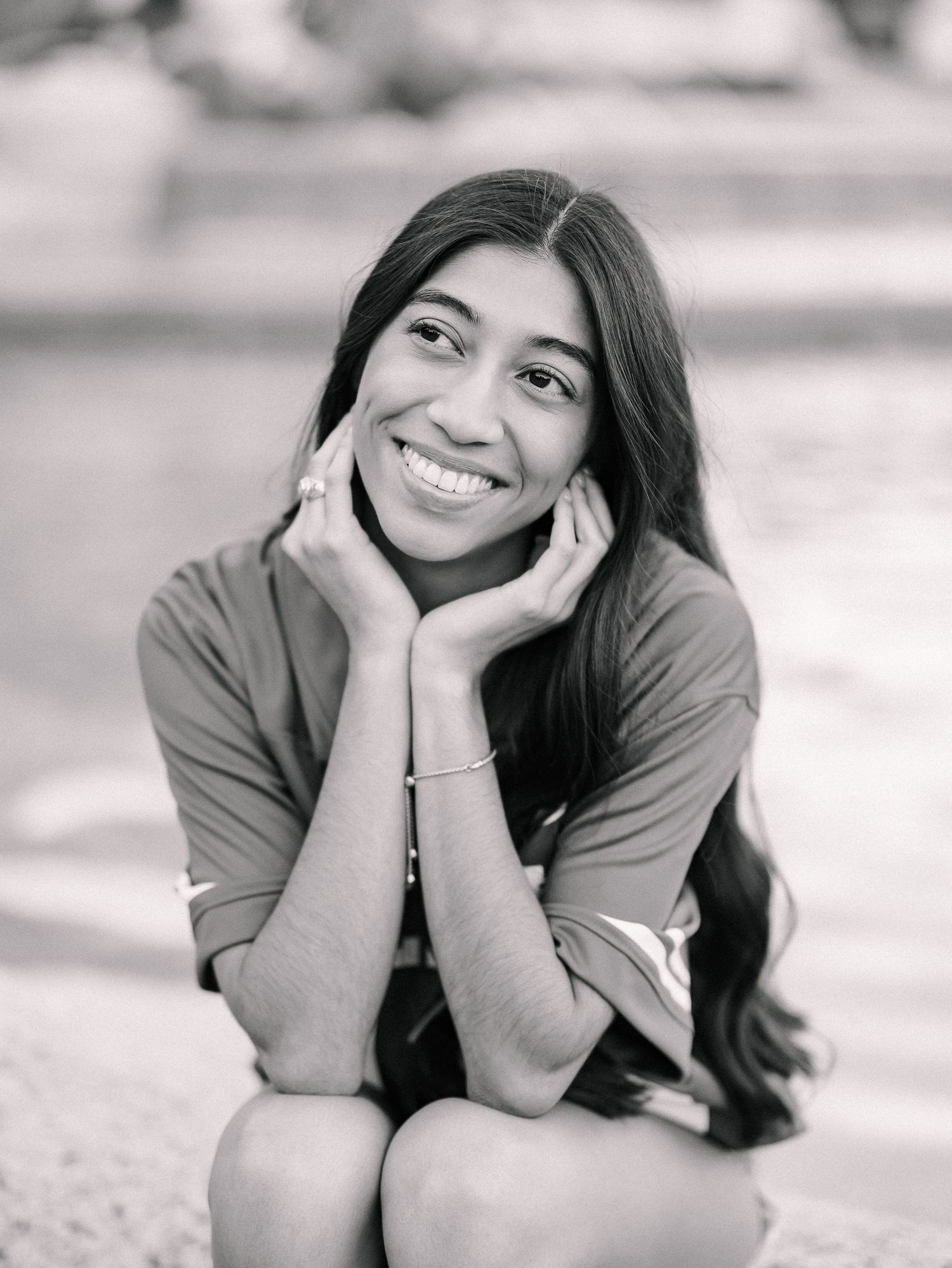 Smiling young woman with long dark hair posing with hands on cheeks in black and white portrait photo.