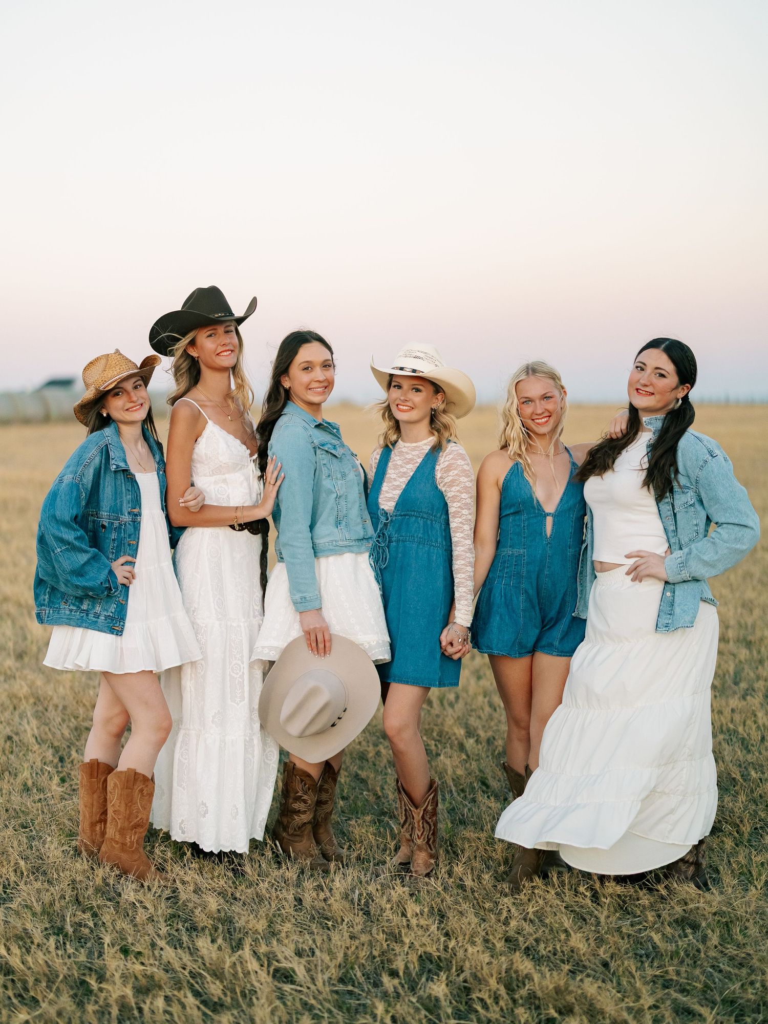 Six women in white dresses, denim jackets, and cowboy hats pose in a golden field at sunset.
