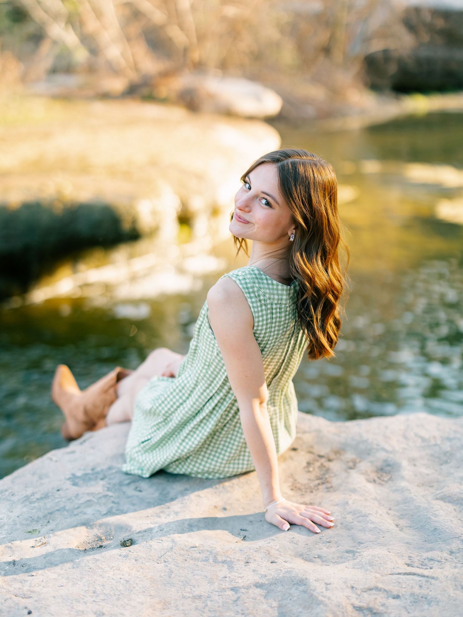 Young woman in green dress and cowboy boots sitting on rocks by a serene creek, gazing upward in golden light.